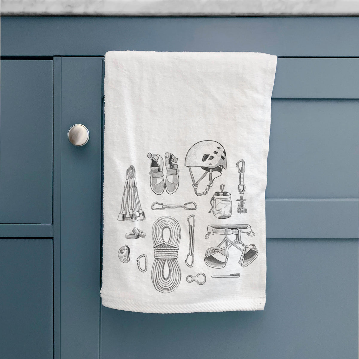White hand towel printed with a black ink flat lay of rock climbing gear, including a helmet, shoes, rope, harness and carabiners, hanging on a cabinet door.