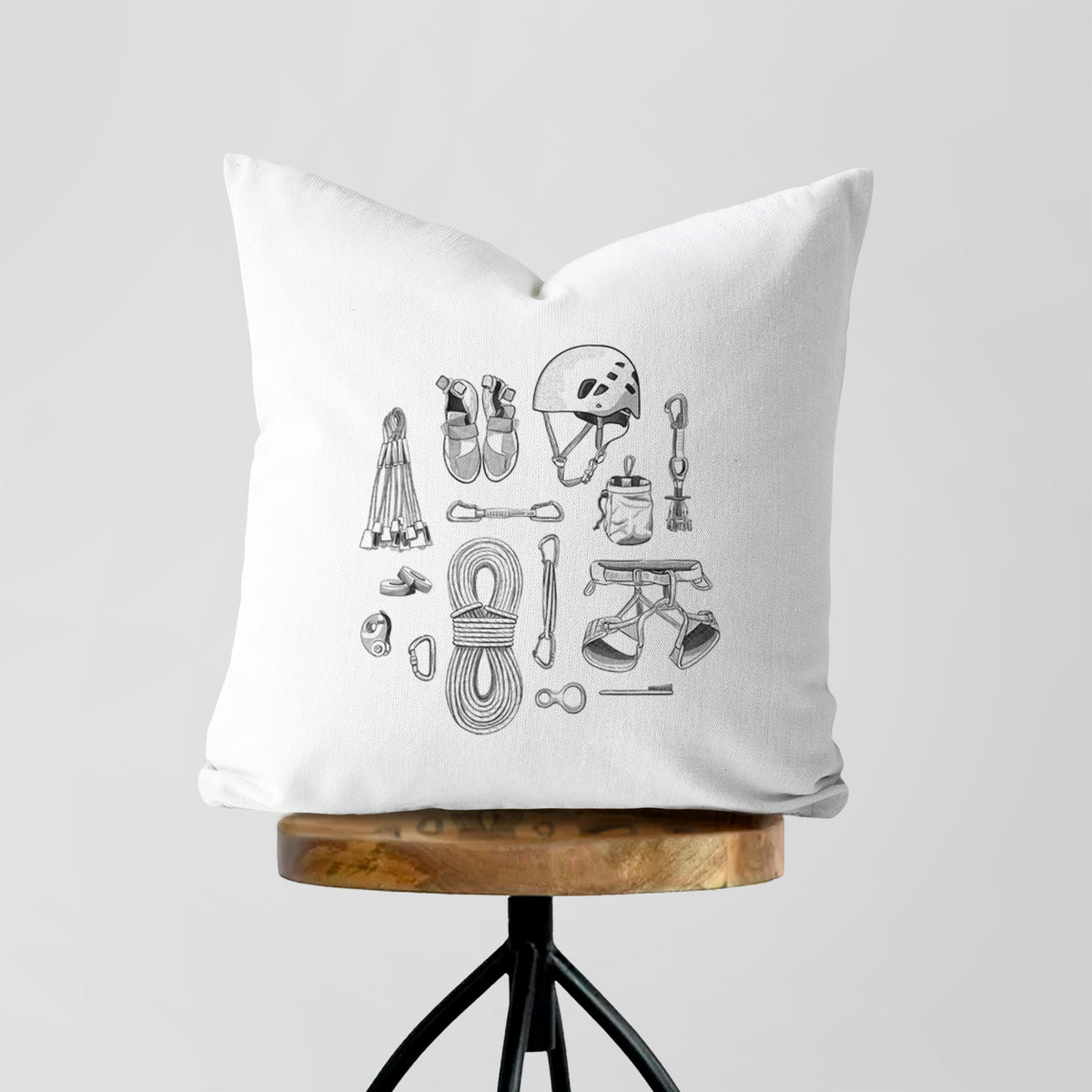 White pillow cover printed with a black ink flat lay of rock climbing gear, including a helmet, shoes, rope, harness and carabiners, on a wooden stool.