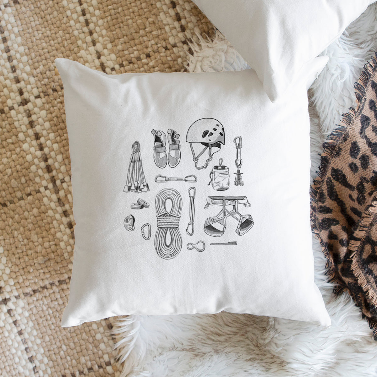 White pillow cover printed with a black ink flat lay of rock climbing gear, including a helmet, shoes, rope, harness and carabiners, on a woven chair.