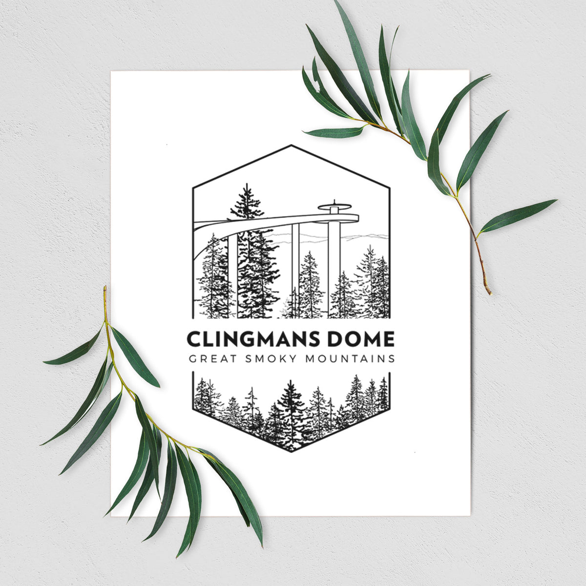 Art print of a black line art badge of the Clingmans Dome observation tower above a pine forest, with the words Clingmans Dome, Great Smoky Mountains, shown with eucalyptus leaves.
