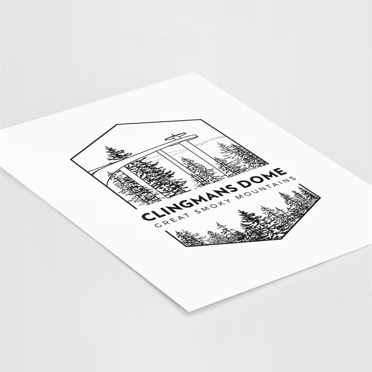 Art print of a black line art badge of the Clingmans Dome observation tower above a pine forest, with the words Clingmans Dome, Great Smoky Mountains, lying at an angle on a white surface.