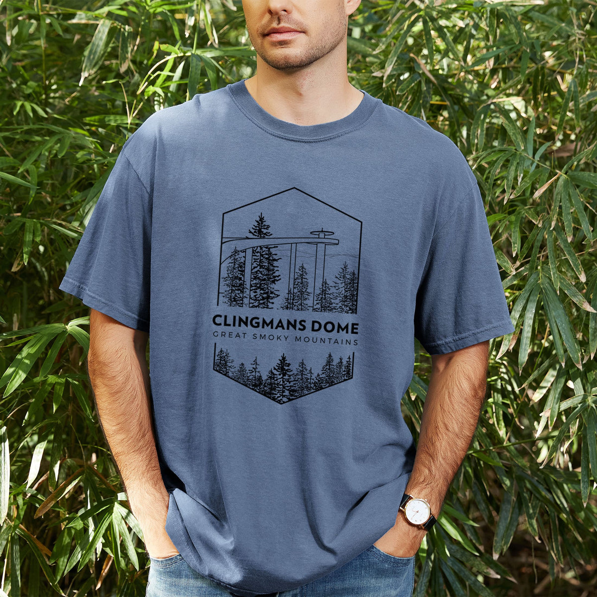 Man wearing a Comfort Colors t-shirt printed with a black line art badge of the Clingmans Dome observation tower above a pine forest, with the words Clingmans Dome, Great Smoky Mountains, outdoors.