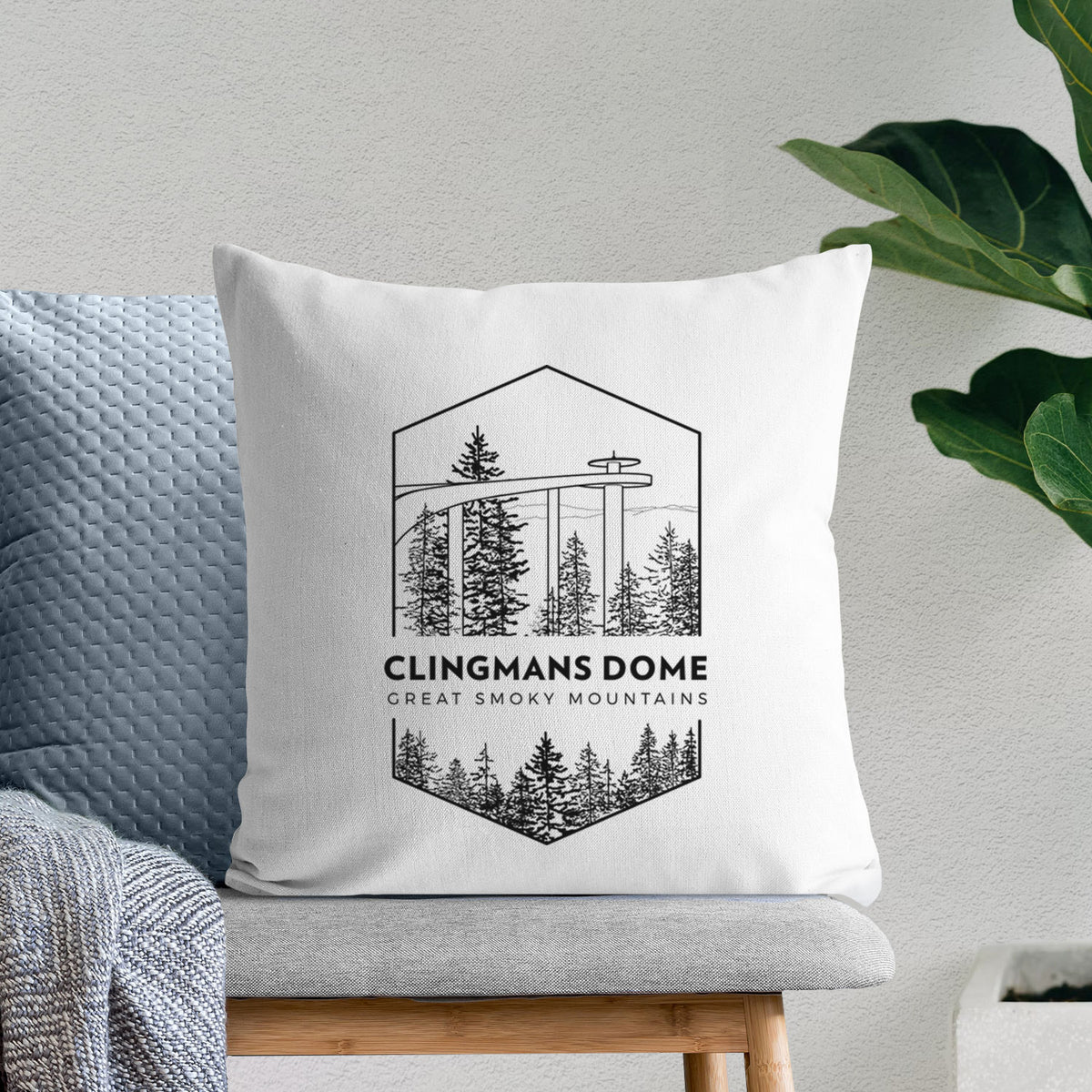 White pillow cover printed with a black line art badge of the Clingmans Dome observation tower above a pine forest, with the words Clingmans Dome, Great Smoky Mountains, on a bench beside plants.