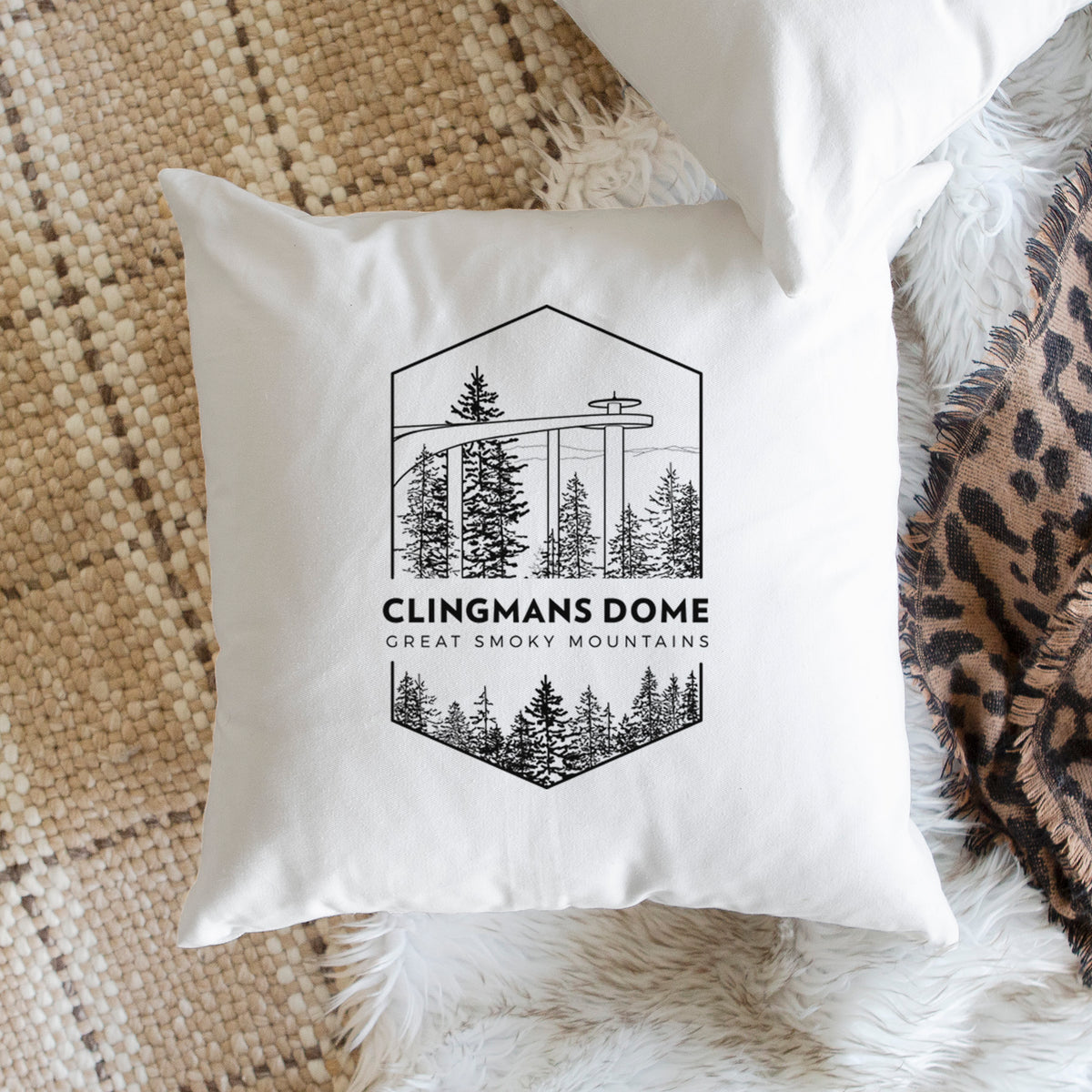 White pillow cover printed with a black line art badge of the Clingmans Dome observation tower above a pine forest, with the words Clingmans Dome, Great Smoky Mountains, on a woven chair.