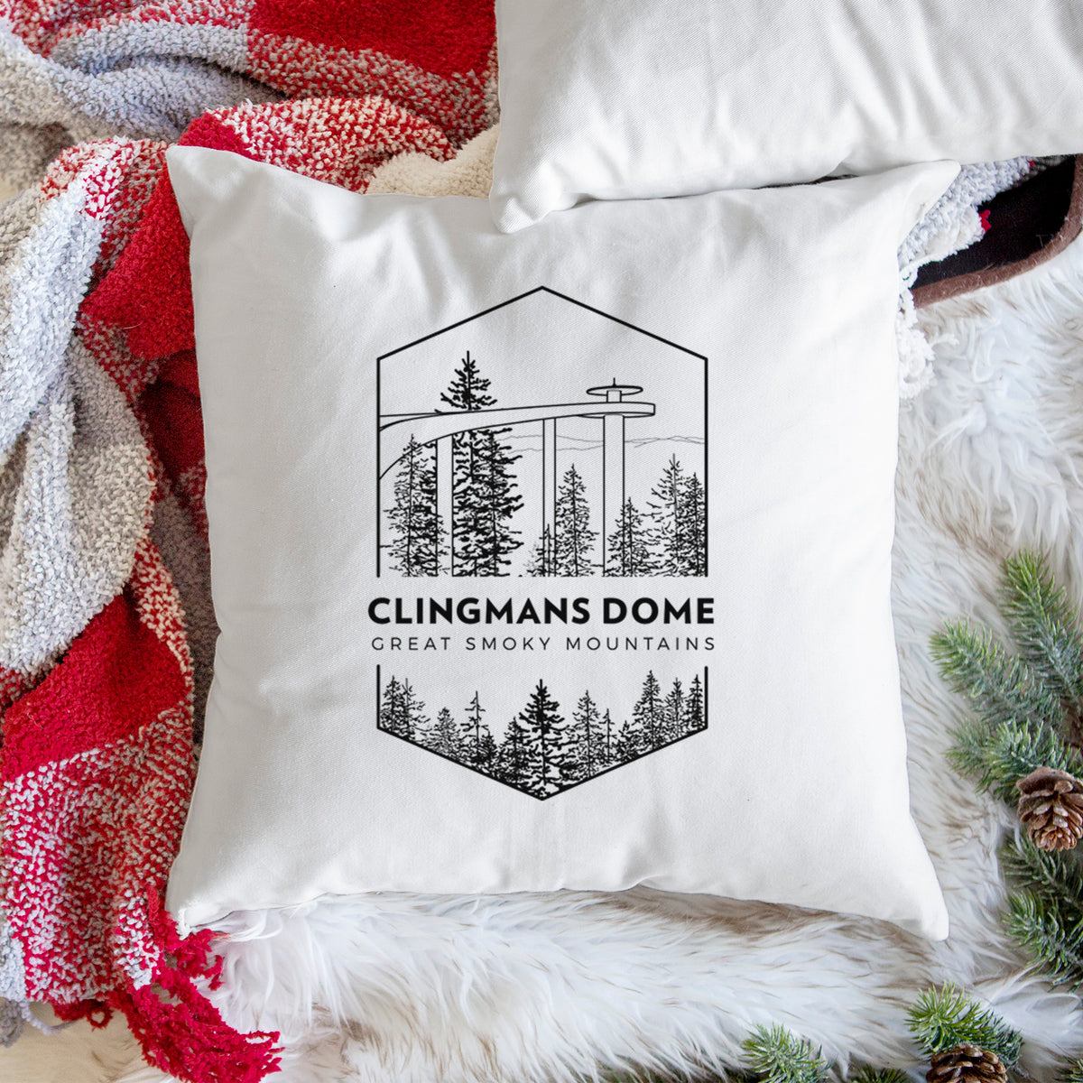 White pillow cover printed with a black line art badge of the Clingmans Dome observation tower above a pine forest, with the words Clingmans Dome, Great Smoky Mountains, on a woven chair.
