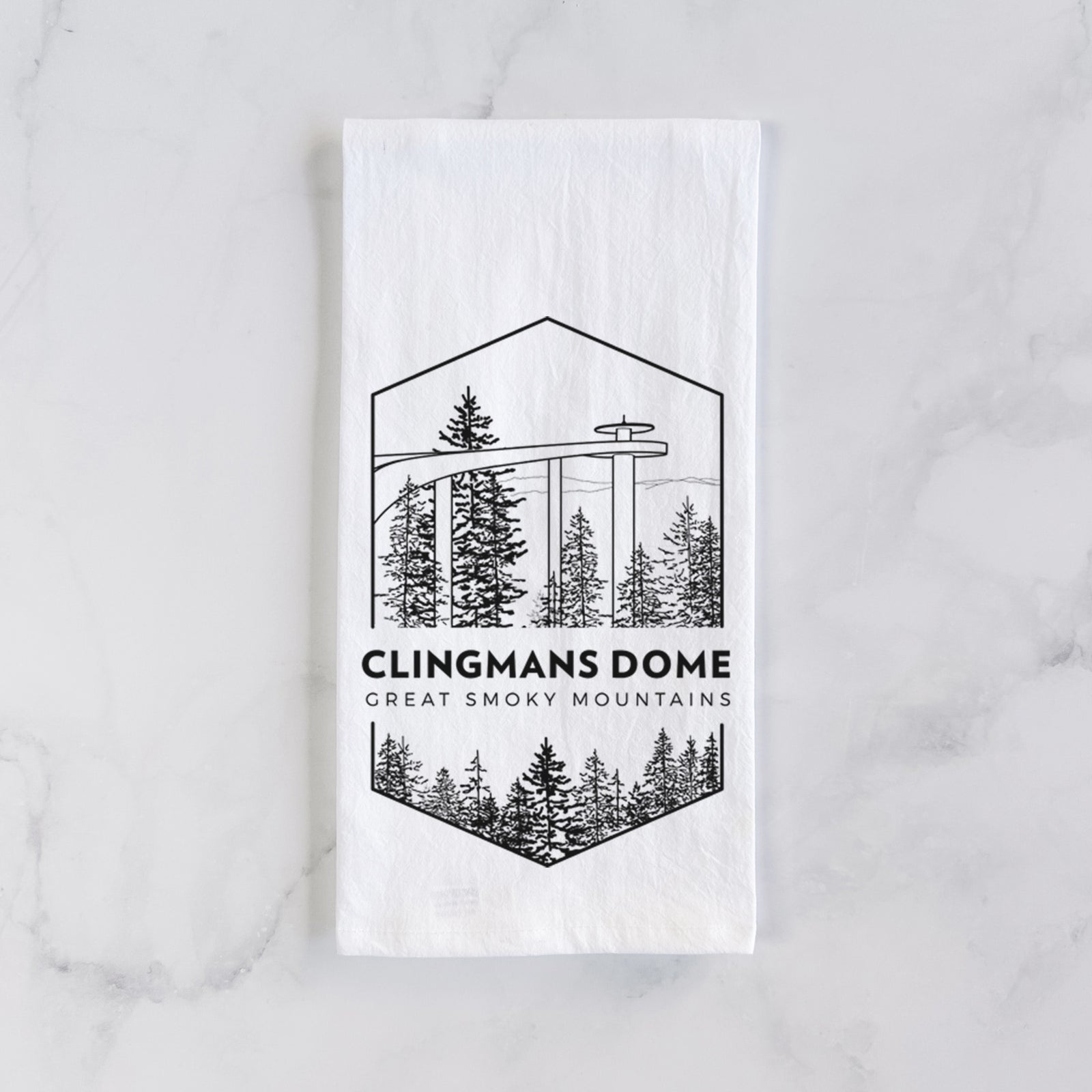 White cotton tea towel printed with a black line art badge of the Clingmans Dome observation tower above a pine forest, with the words Clingmans Dome, Great Smoky Mountains, on a marble counter beside a round wooden cutting board and greenery.