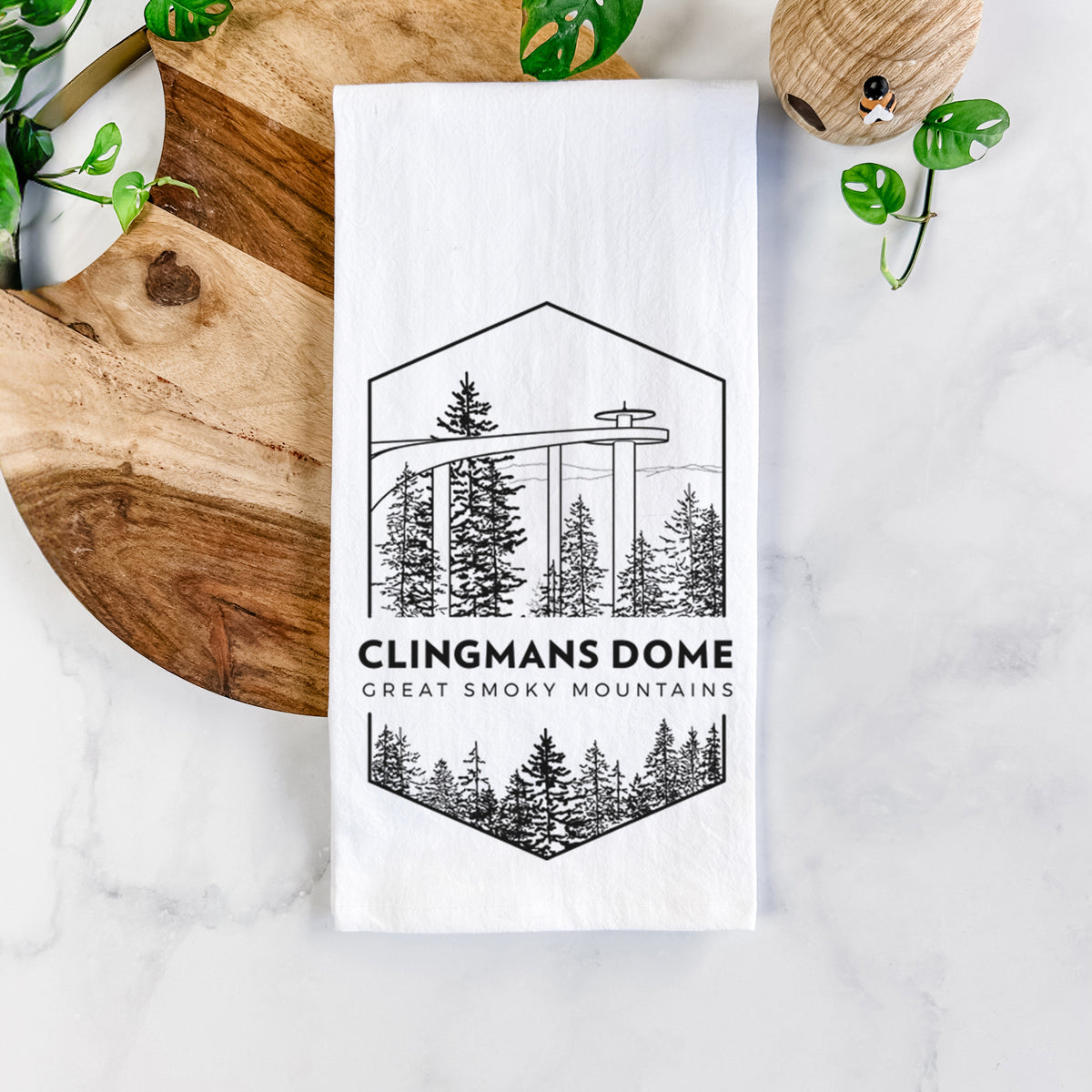 White cotton tea towel printed with a black line art badge of the Clingmans Dome observation tower above a pine forest, with the words Clingmans Dome, Great Smoky Mountains, on a marble counter beside a round wooden cutting board and greenery.