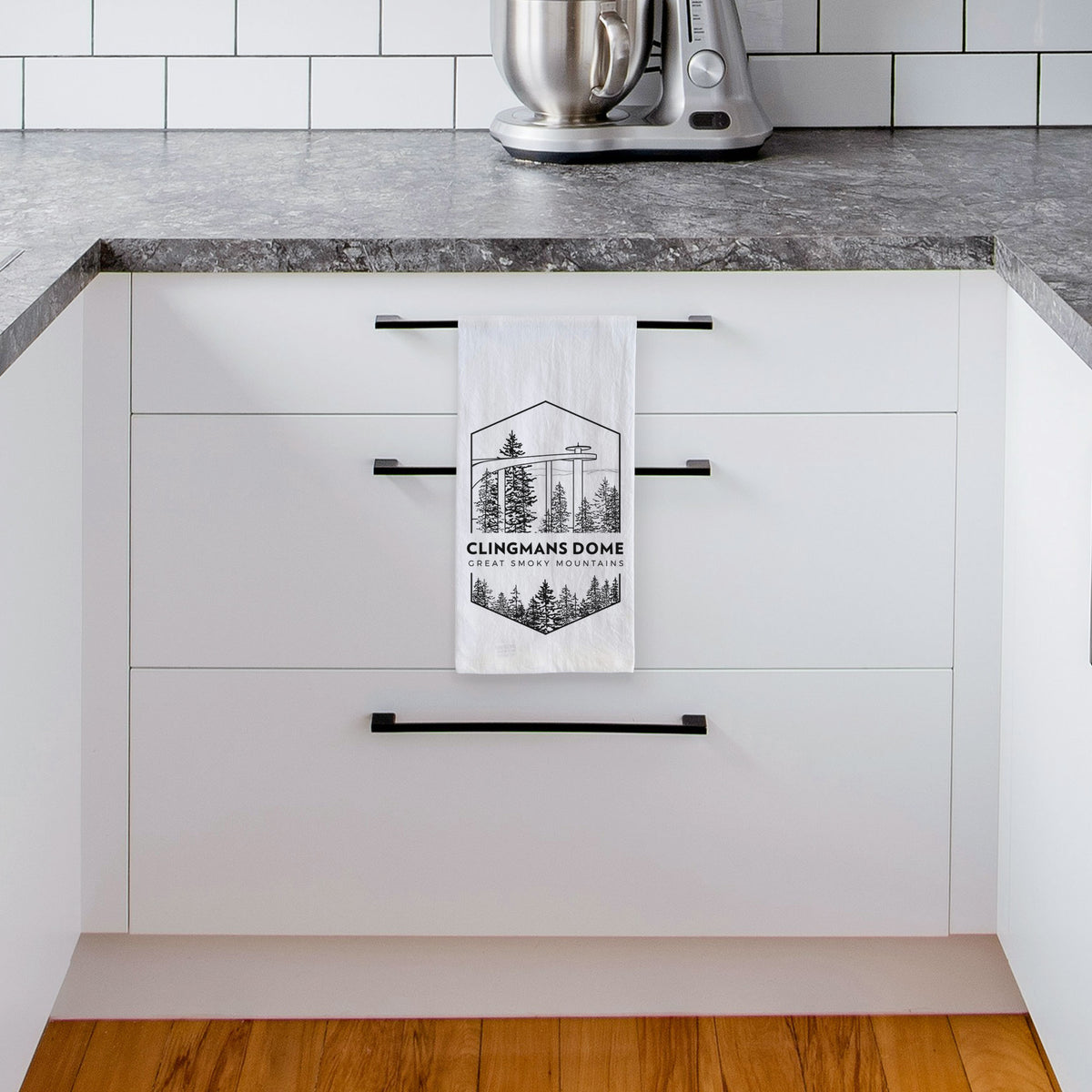 White tea towel printed with a black line art badge of the Clingmans Dome observation tower above a pine forest, with the words Clingmans Dome, Great Smoky Mountains, hanging from a drawer handle in a bright white kitchen.