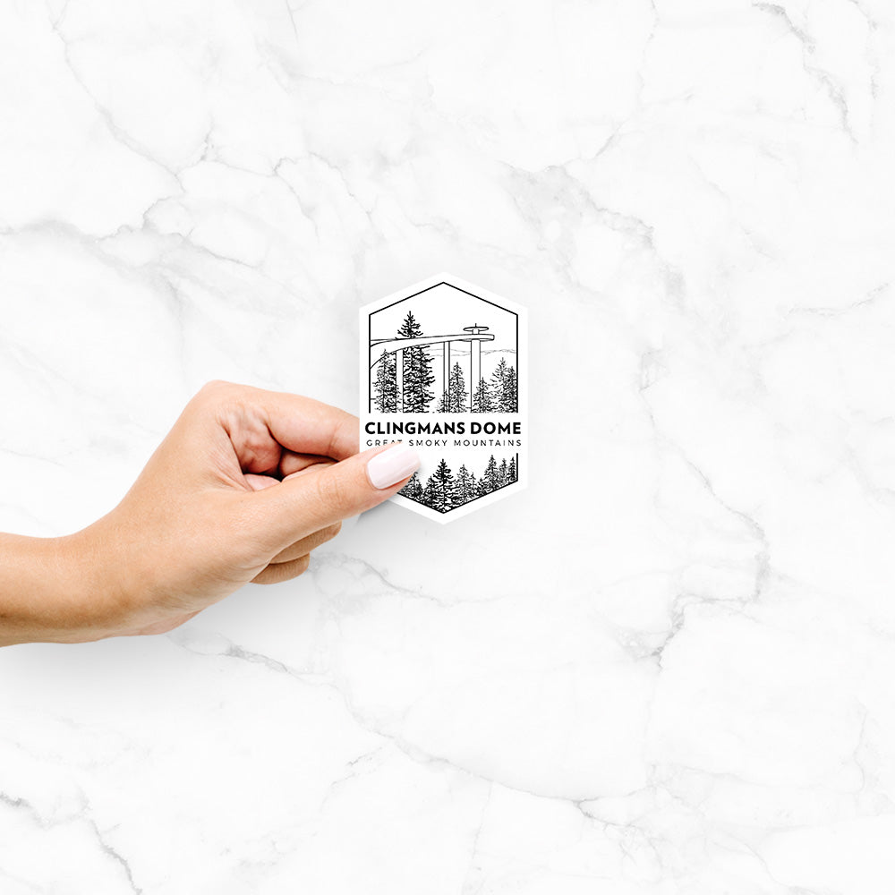 Hand holding a 4 inch vinyl decal of a black line art badge of the Clingmans Dome observation tower above a pine forest, with the words Clingmans Dome, Great Smoky Mountains.