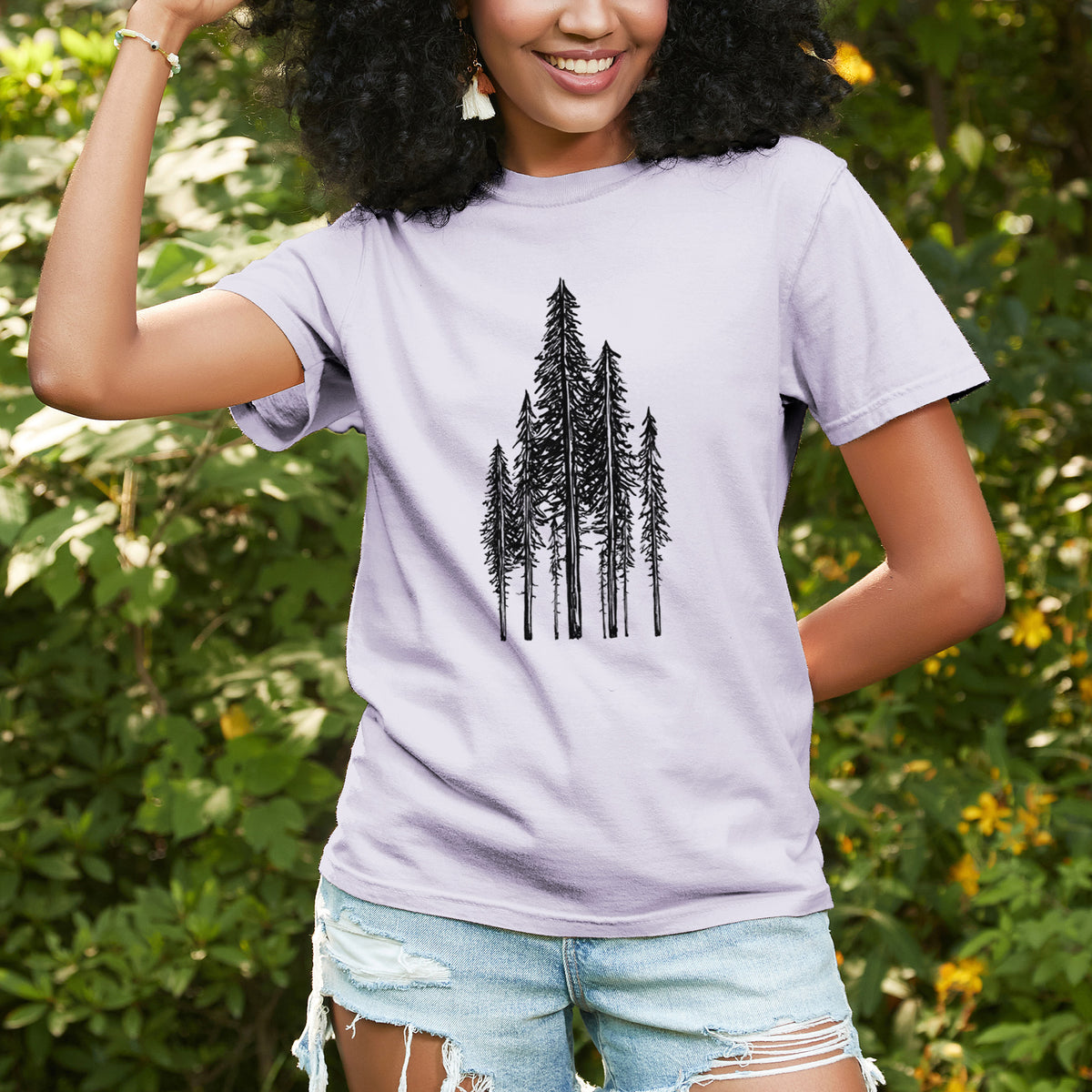 Woman wearing a Comfort Colors t-shirt printed with a black ink illustration of a stand of tall coastal redwood trees with slender trunks and layered branches, outdoors.
