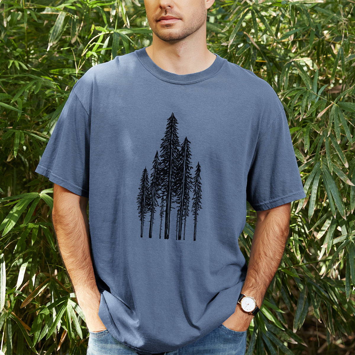 Man wearing a Comfort Colors t-shirt printed with a black ink illustration of a stand of tall coastal redwood trees with slender trunks and layered branches, outdoors.
