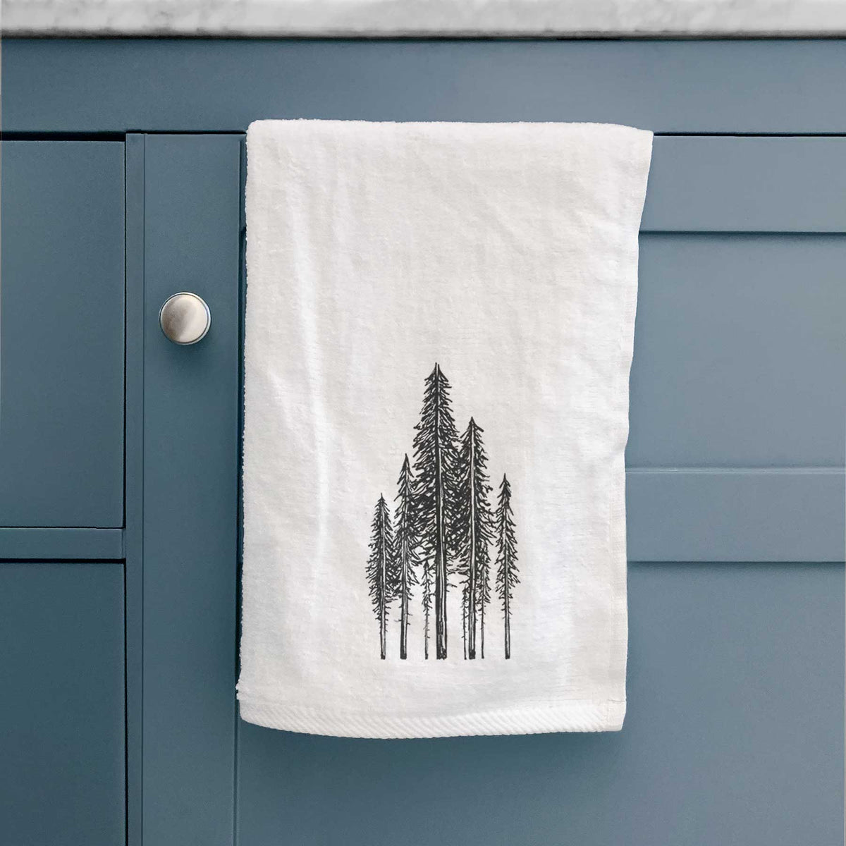 White hand towel printed with a black ink illustration of a stand of tall coastal redwood trees with slender trunks and layered branches, hanging on a cabinet door.