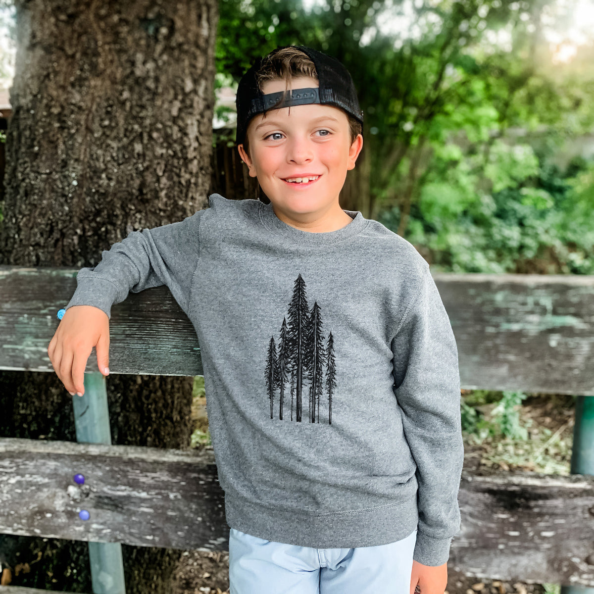 Boy wearing a youth crewneck sweatshirt printed with a black ink illustration of a stand of tall coastal redwood trees with slender trunks and layered branches, outdoors.