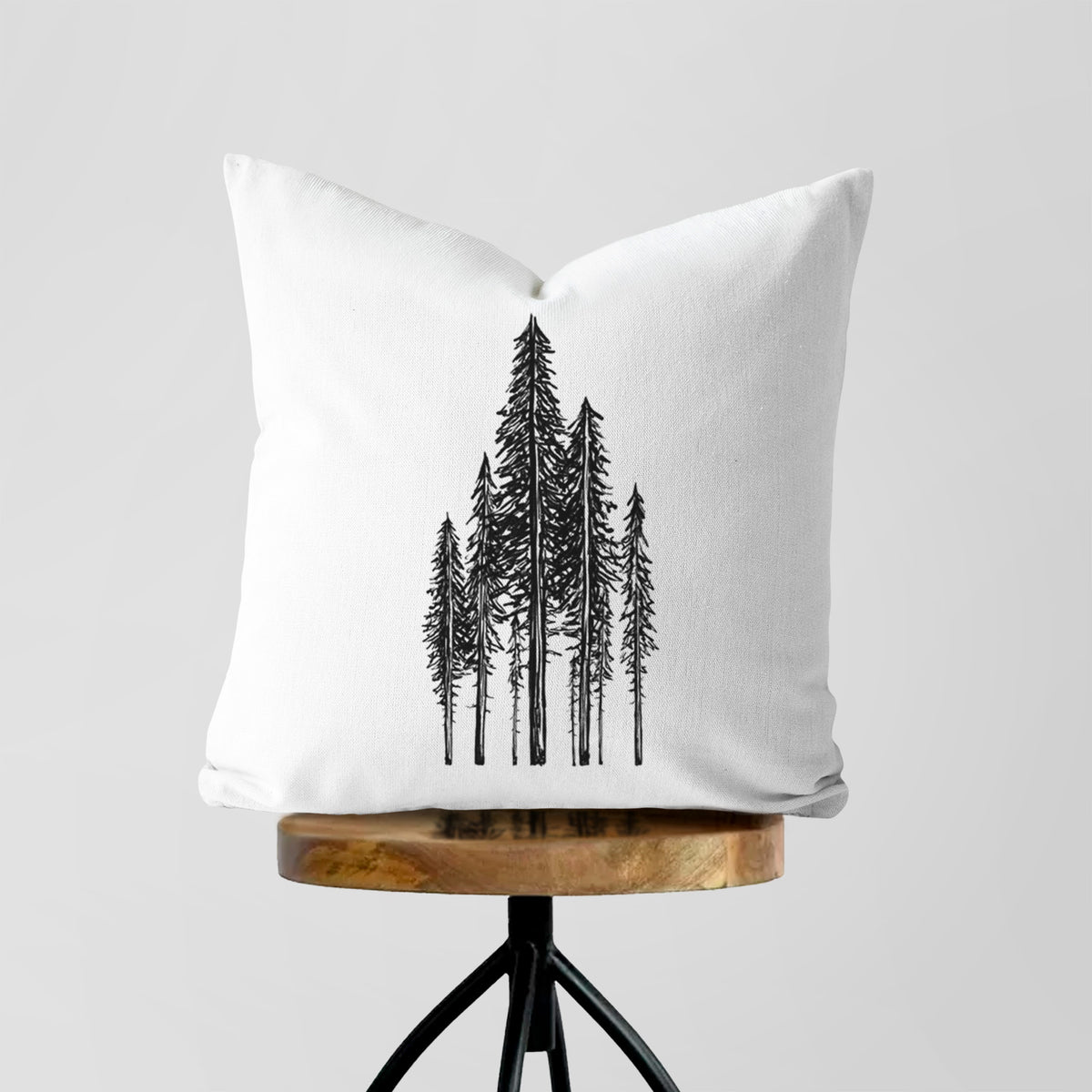 White pillow cover printed with a black ink illustration of a stand of tall coastal redwood trees with slender trunks and layered branches, on a wooden stool.
