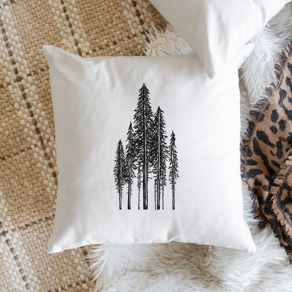 White pillow cover printed with a black ink illustration of a stand of tall coastal redwood trees with slender trunks and layered branches, on a woven chair.