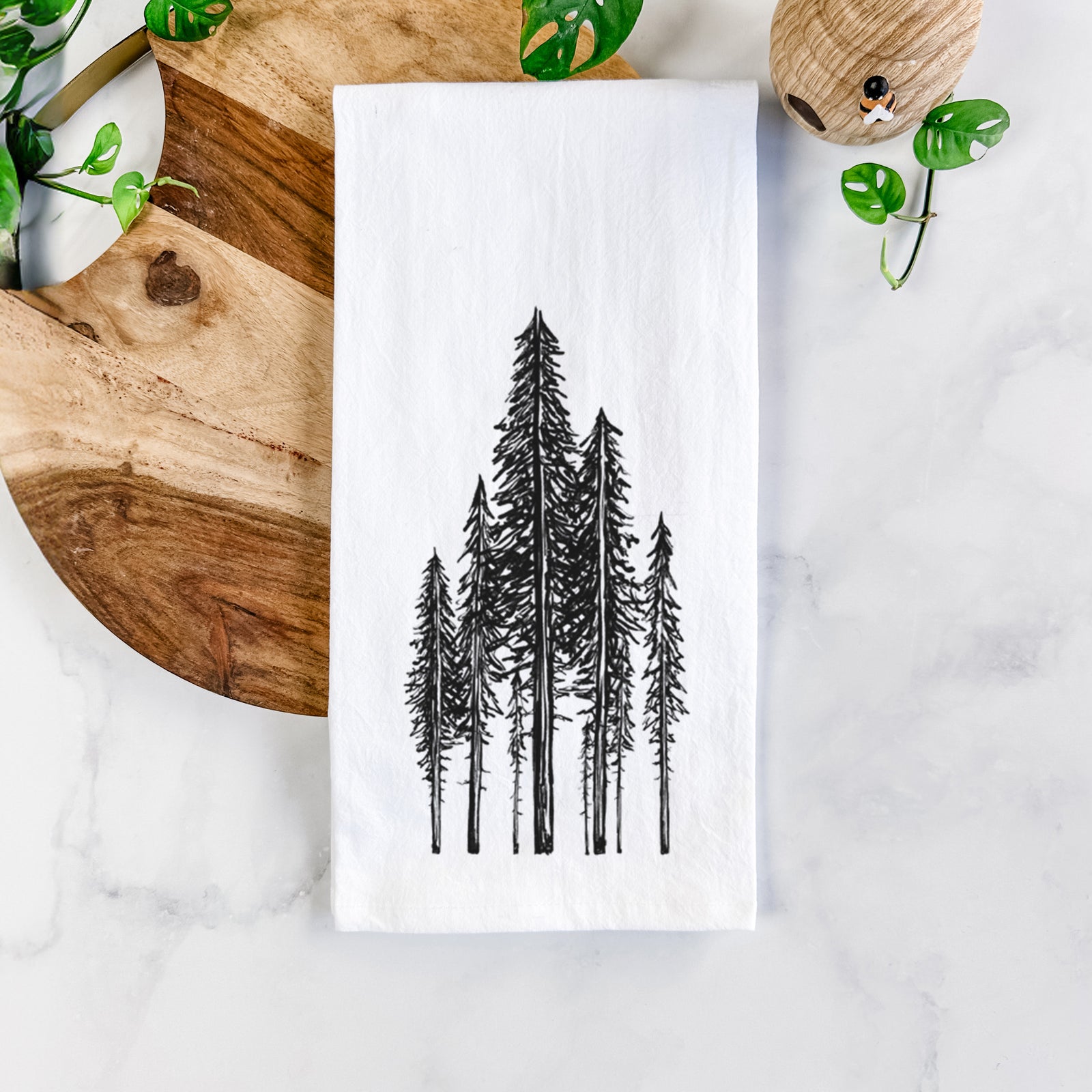 White cotton tea towel printed with a black ink illustration of a stand of tall coastal redwood trees with slender trunks and layered branches, on a marble counter beside a round wooden cutting board and greenery.