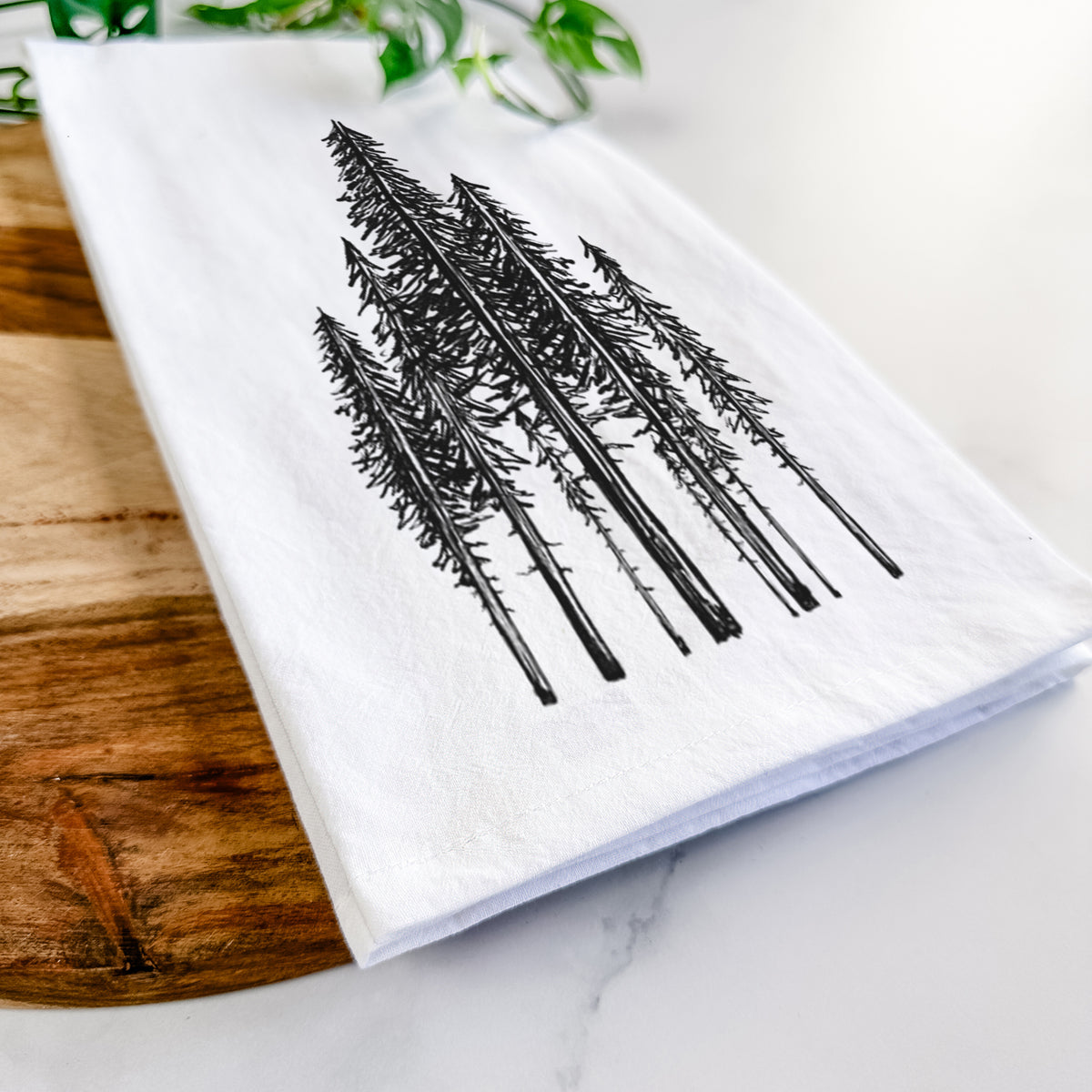 Close-up of a folded white tea towel on a wooden board, showing a black ink illustration of a stand of tall coastal redwood trees with slender trunks and layered branches.