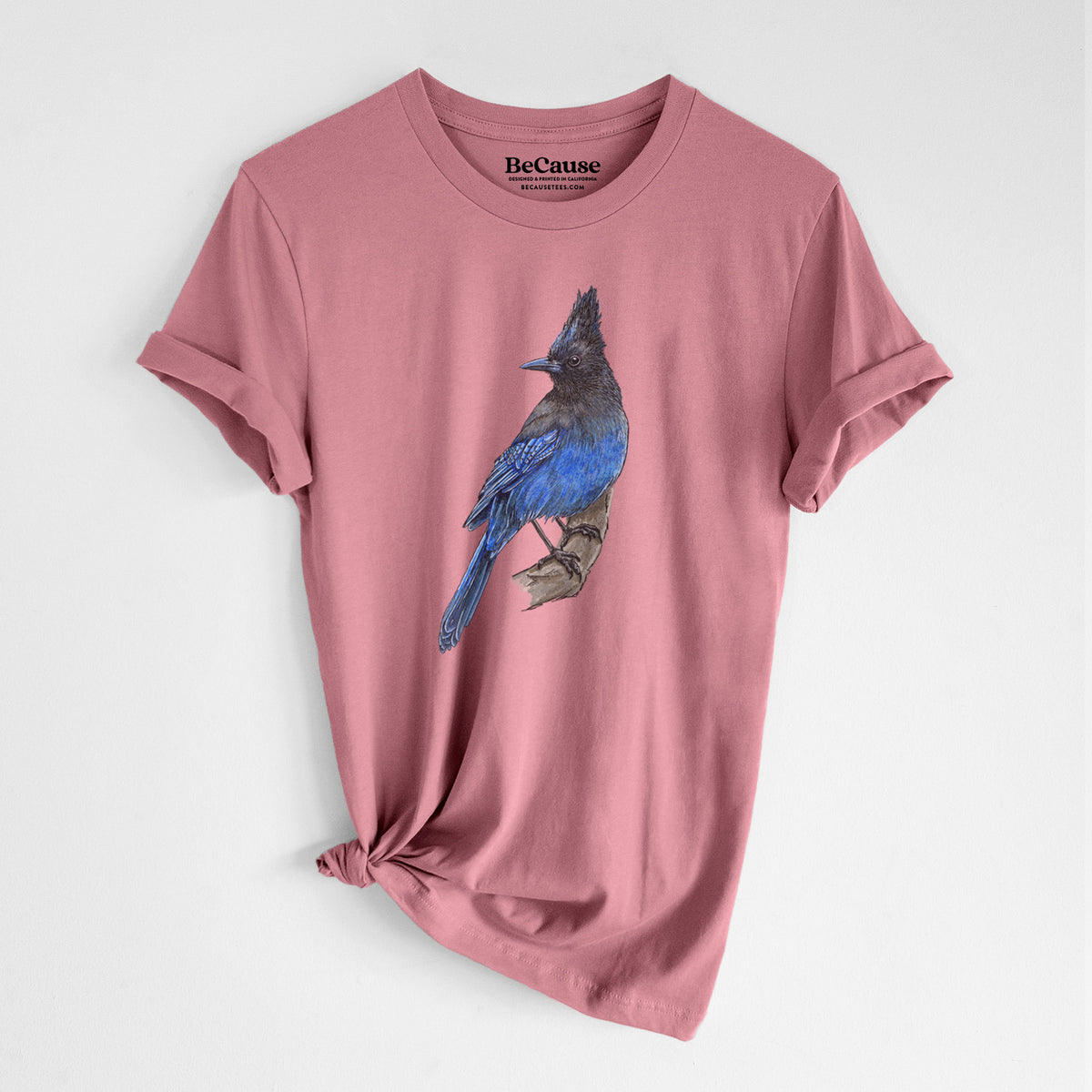 Mauve pink lightweight cotton t-shirt laid flat with the sleeves rolled, printed with a full-color illustration of a Steller's jay perched with its dark crest raised.
