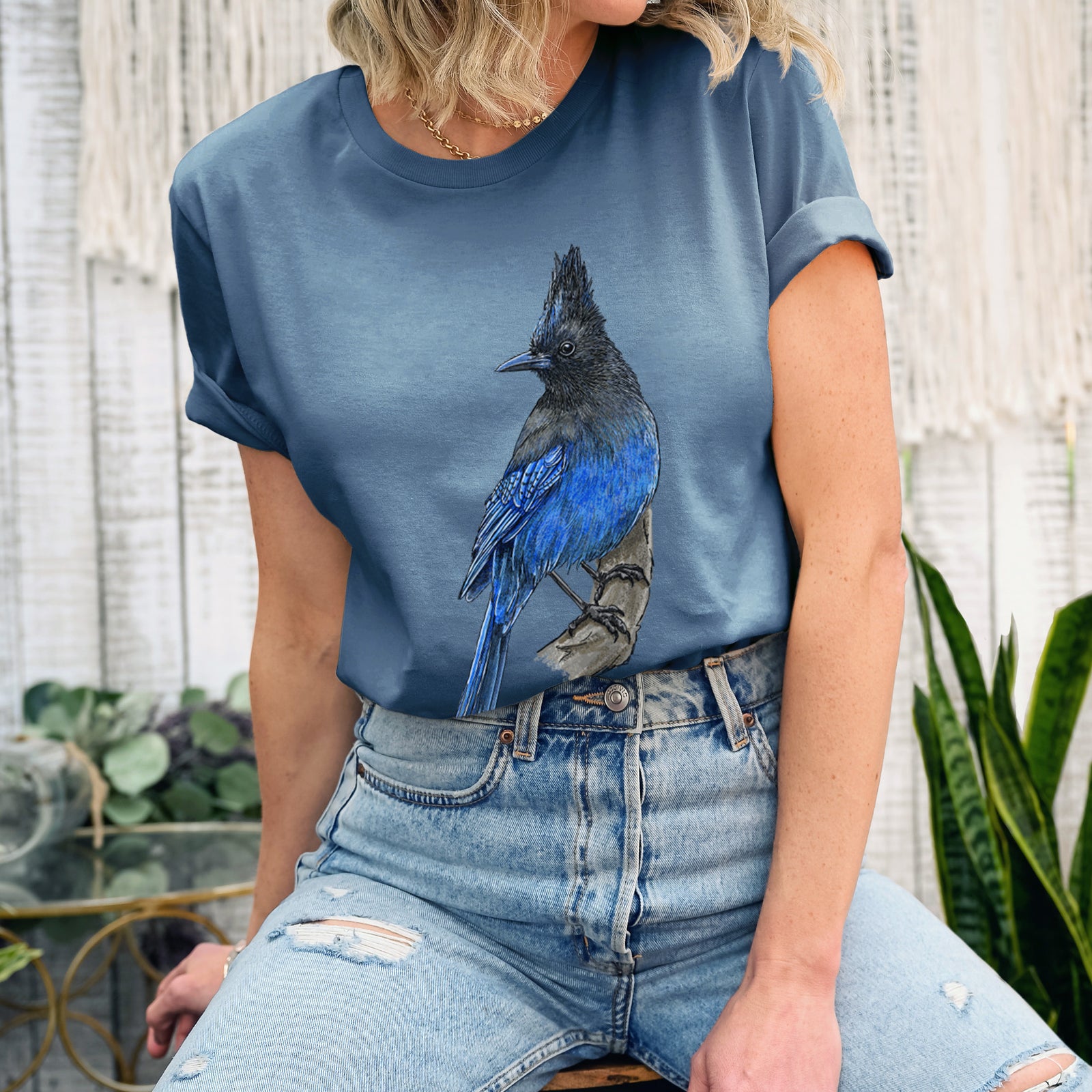 Autumn lightweight cotton t-shirt laid flat, printed with a full-color illustration of a Steller's jay perched with its dark crest raised.