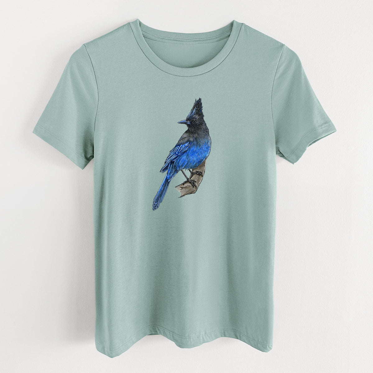 Dusty blue women's relaxed fit cotton t-shirt laid flat, printed with a full-color illustration of a Steller's jay perched with its dark crest raised.