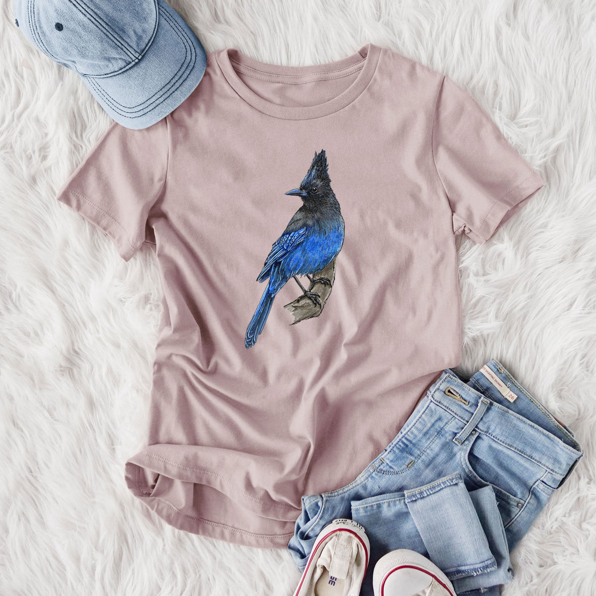 Women's relaxed fit cotton t-shirt printed with a full-color illustration of a Steller's jay perched with its dark crest raised, styled flat on a white faux fur rug with jeans.