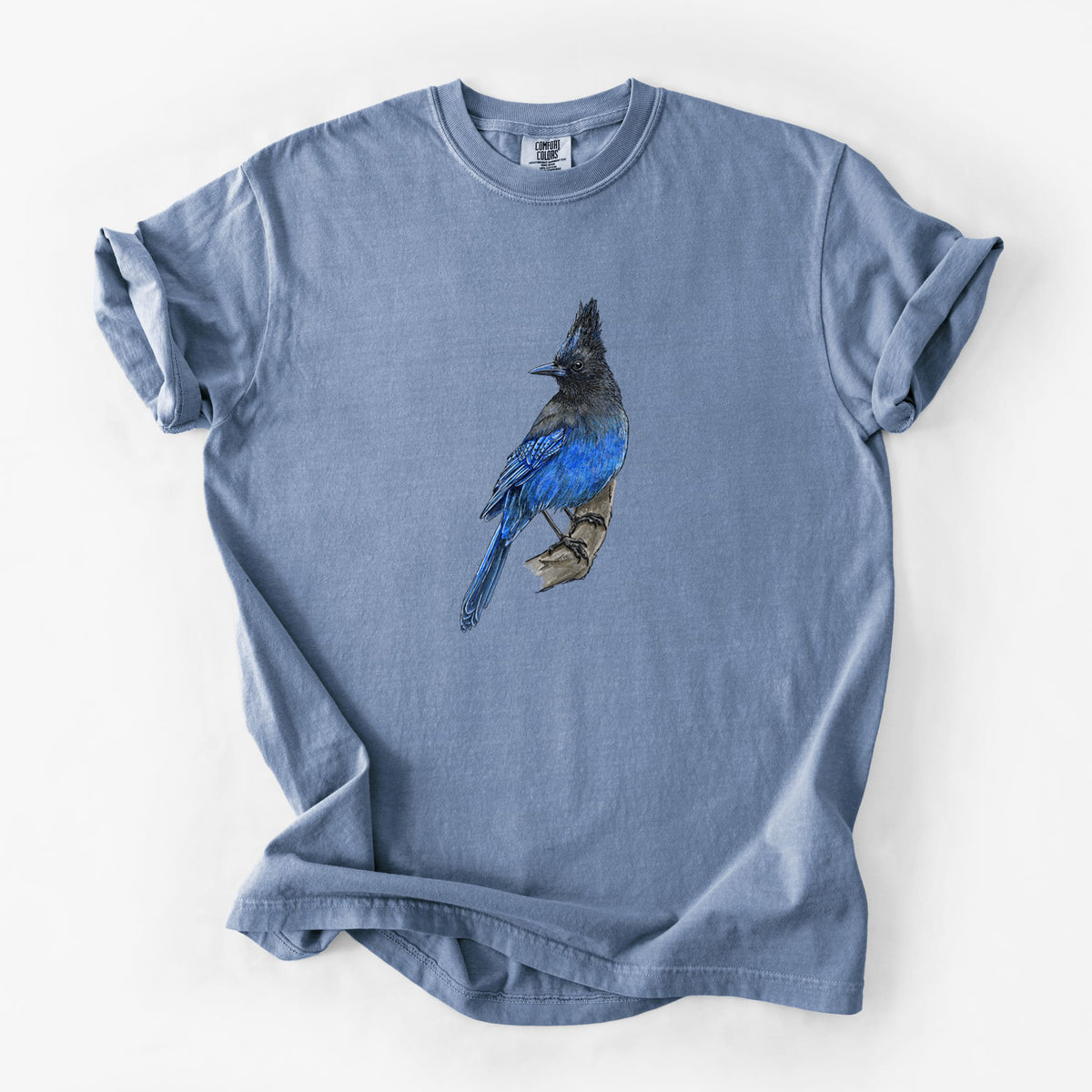 Blue jean Comfort Colors t-shirt laid flat, printed with a full-color illustration of a Steller's jay perched with its dark crest raised.