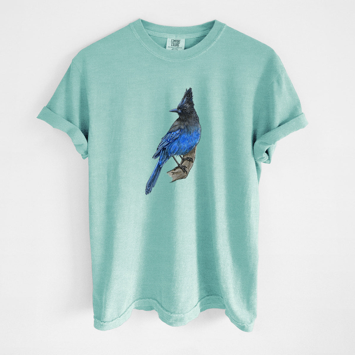Chalky mint Comfort Colors t-shirt laid flat, printed with a full-color illustration of a Steller's jay perched with its dark crest raised.