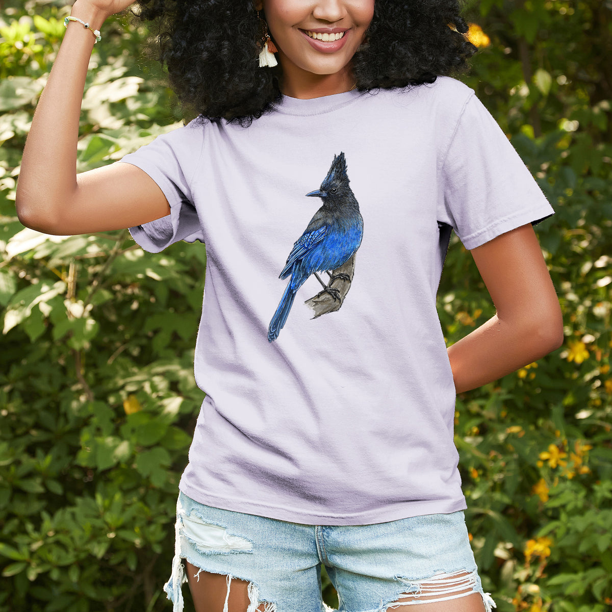 Woman wearing a Comfort Colors t-shirt printed with a full-color illustration of a Steller's jay perched with its dark crest raised, outdoors.