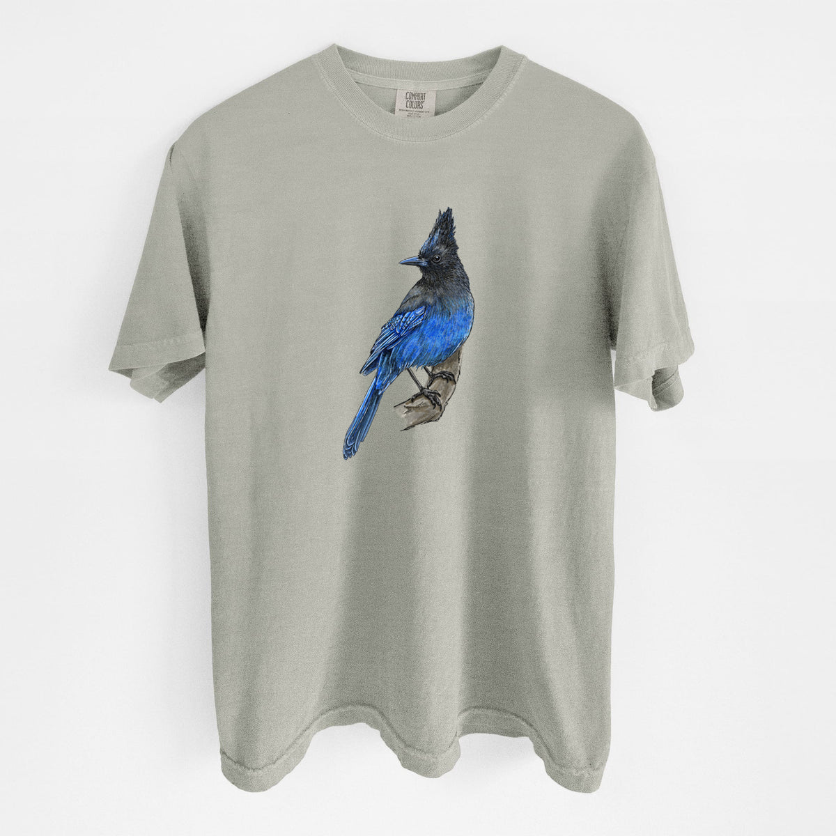 Sandstone beige Comfort Colors t-shirt laid flat, printed with a full-color illustration of a Steller's jay perched with its dark crest raised.