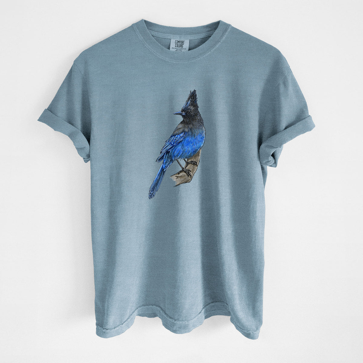 Steel blue Comfort Colors t-shirt laid flat, printed with a full-color illustration of a Steller's jay perched with its dark crest raised.