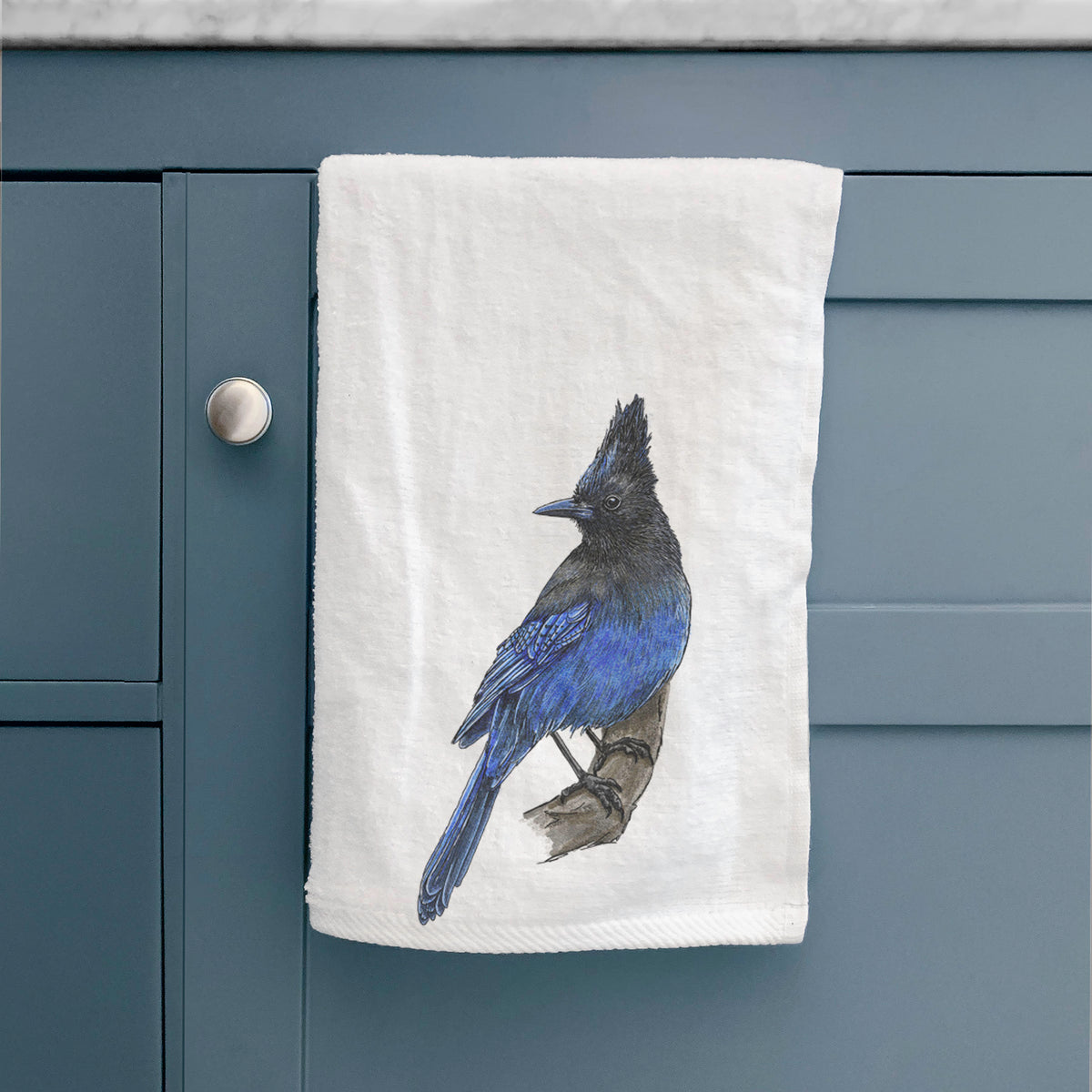 White hand towel printed with a full-color illustration of a Steller's jay perched with its dark crest raised, hanging on a cabinet door.