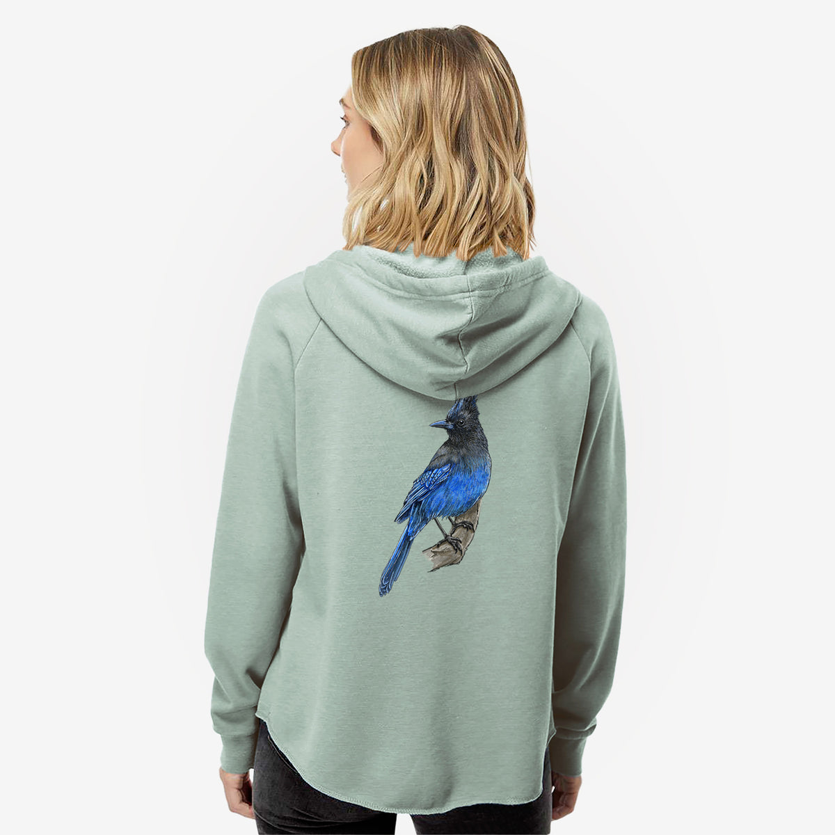 Woman wearing a sage green women's Cali Wave zip-up sweatshirt printed with a full-color illustration of a Steller's jay perched with its dark crest raised, seen from behind.