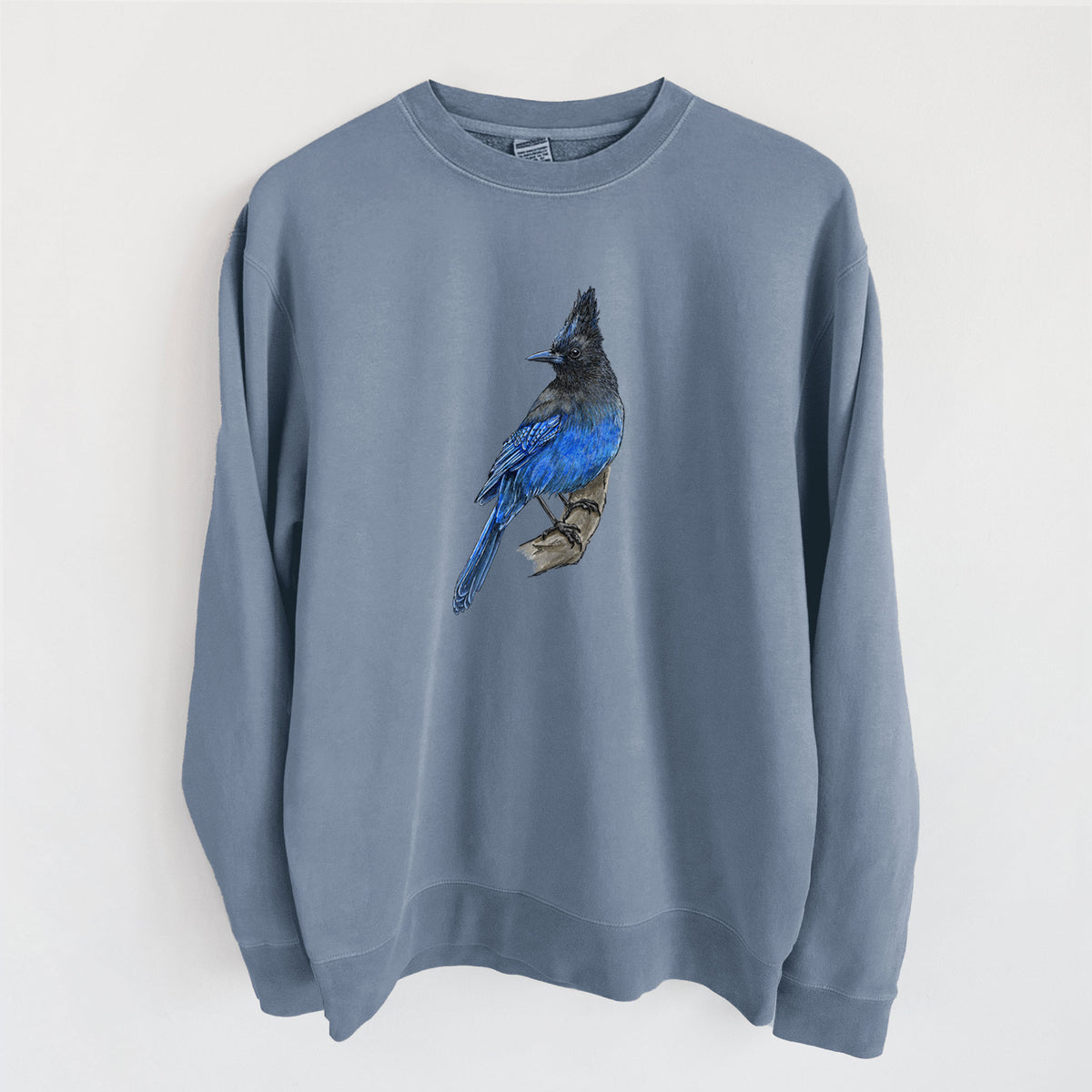 Slate blue pigment dyed crewneck sweatshirt laid flat, printed with a full-color illustration of a Steller's jay perched with its dark crest raised.