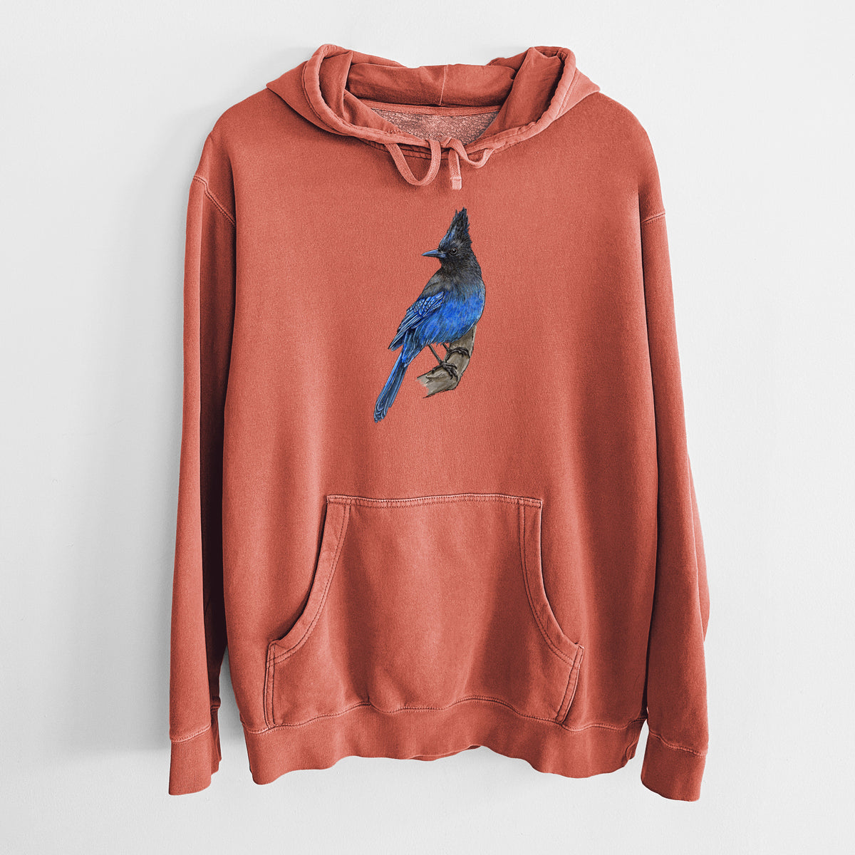 Amber orange pigment dyed hoodie laid flat, printed with a full-color illustration of a Steller's jay perched with its dark crest raised.
