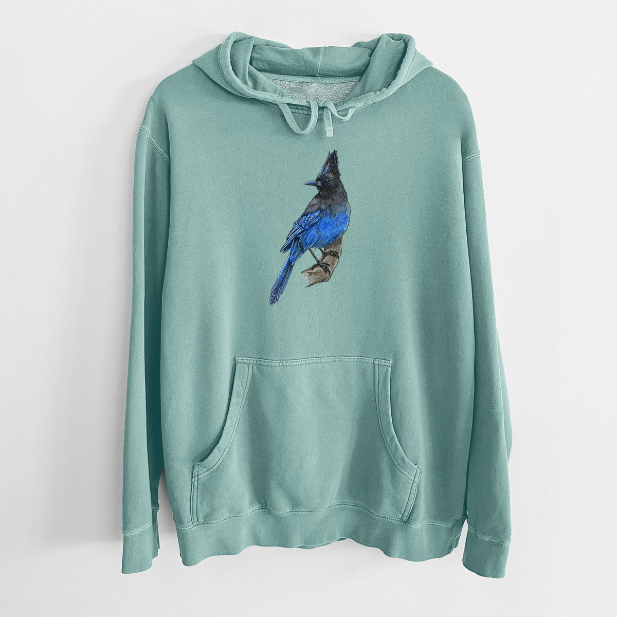 Mint green pigment dyed hoodie laid flat, printed with a full-color illustration of a Steller's jay perched with its dark crest raised.