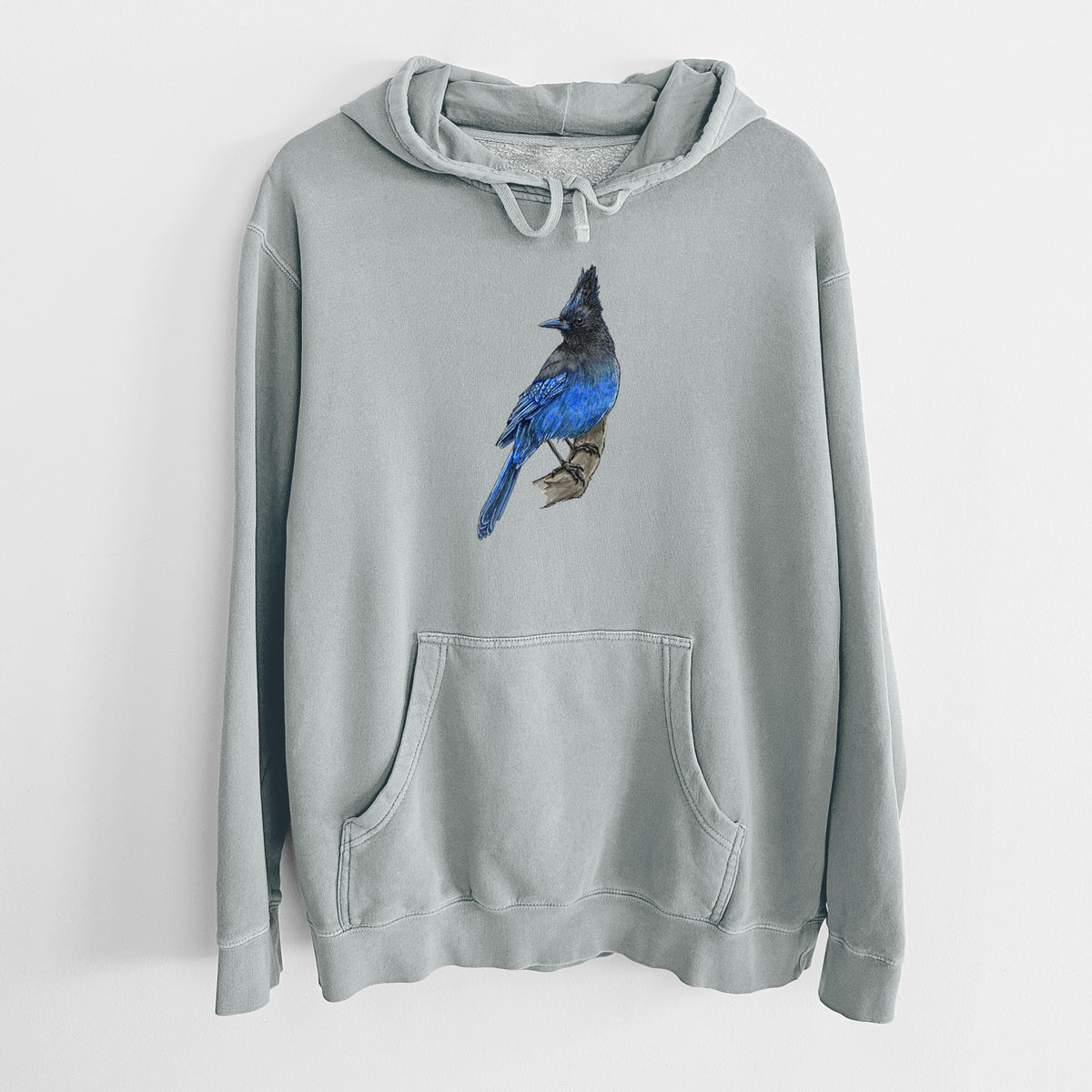 Sage green pigment dyed hoodie laid flat, printed with a full-color illustration of a Steller's jay perched with its dark crest raised.