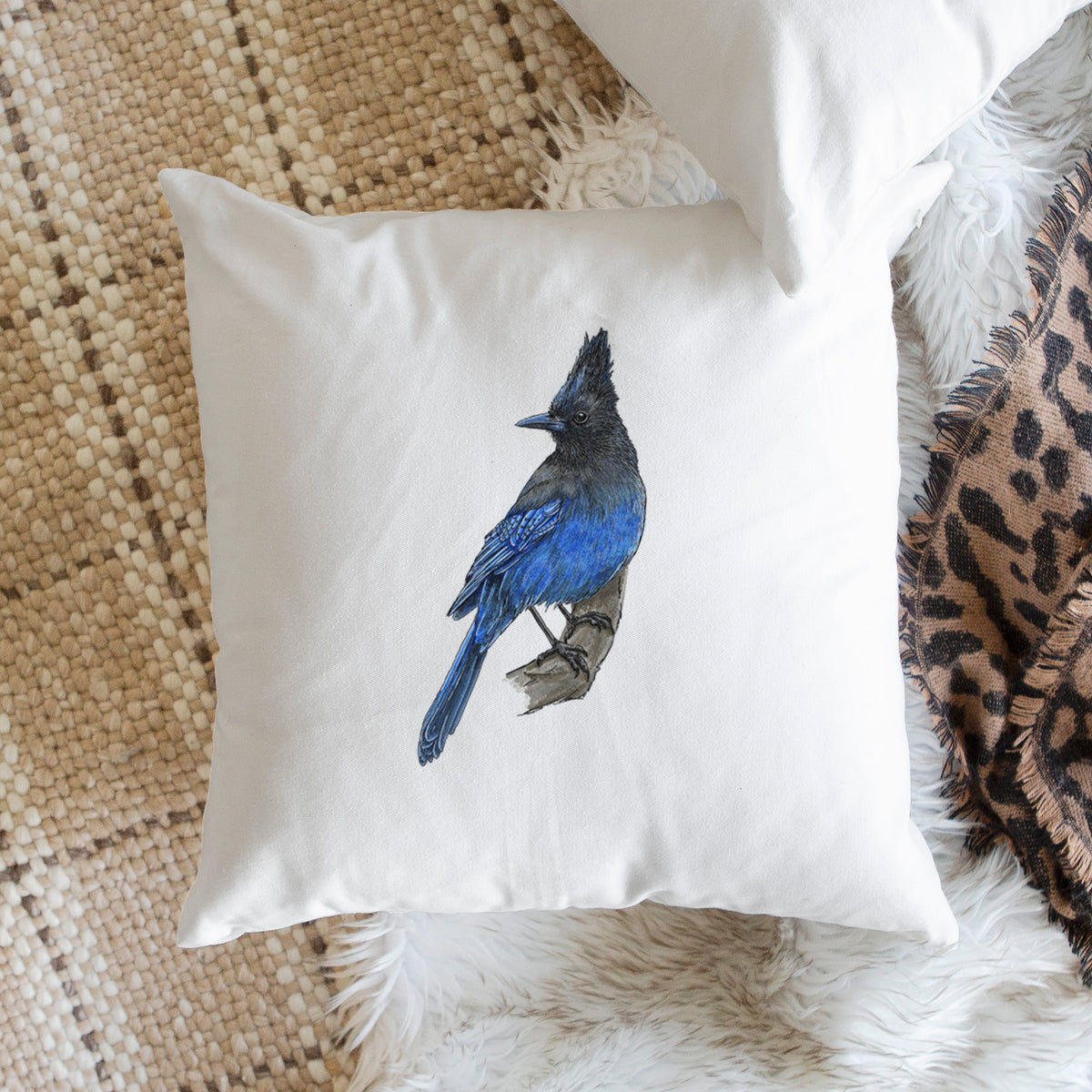 White pillow cover printed with a full-color illustration of a Steller's jay perched with its dark crest raised, on a woven chair.