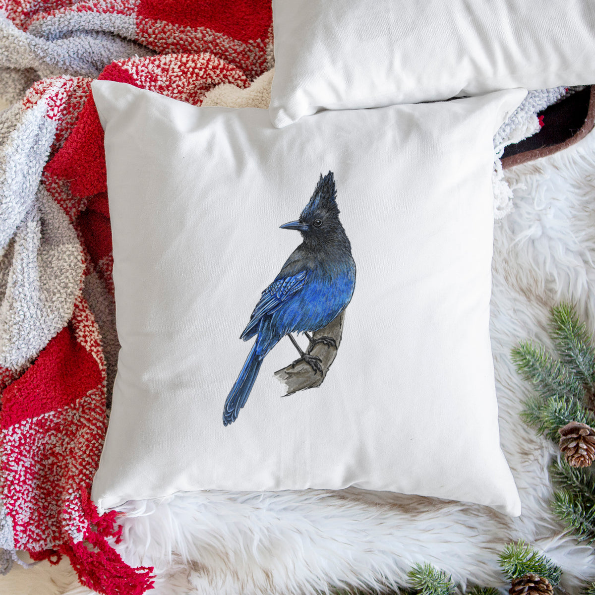 White pillow cover printed with a full-color illustration of a Steller's jay perched with its dark crest raised, on a woven chair.