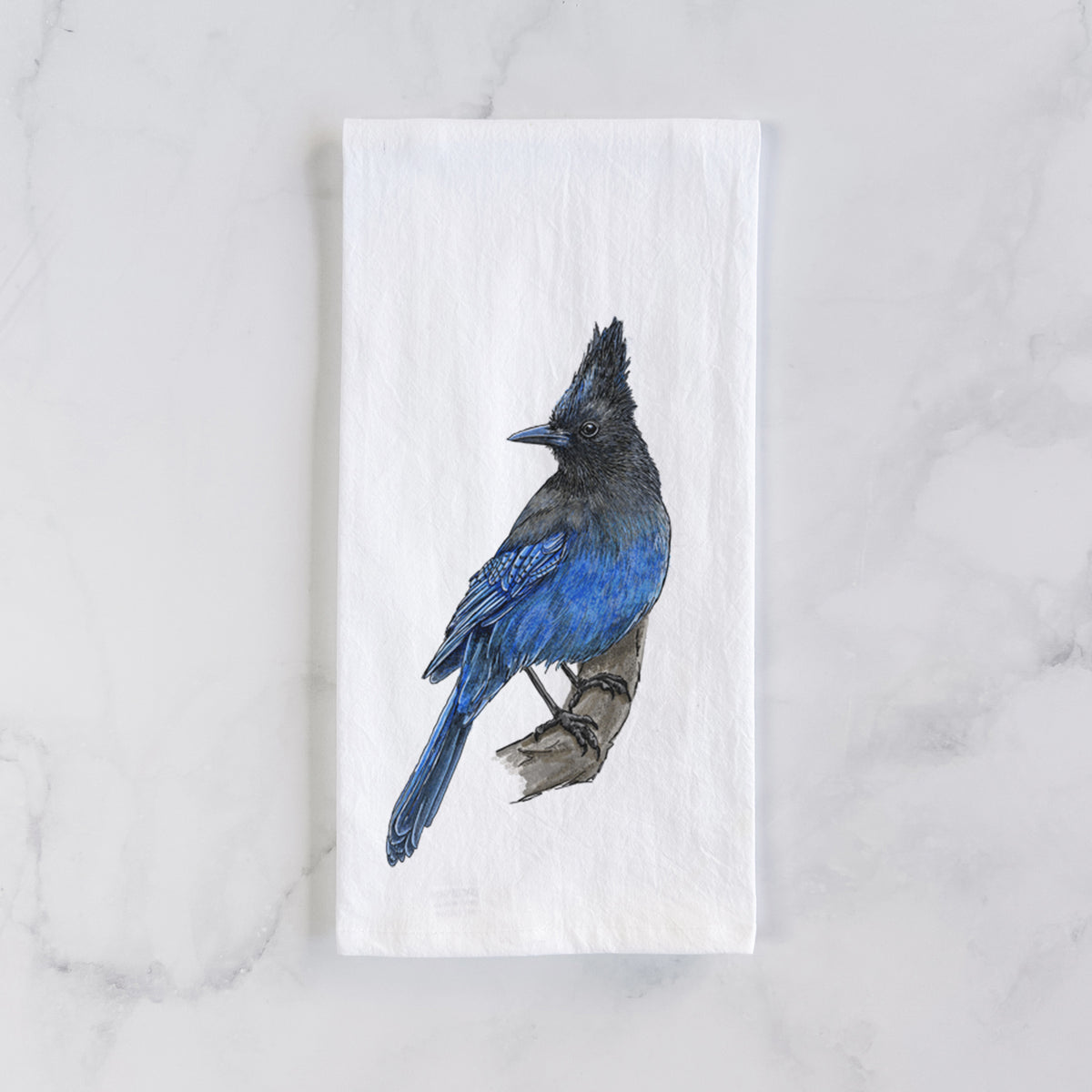 White cotton tea towel laid flat on marble, printed with a full-color illustration of a Steller's jay perched with its dark crest raised.