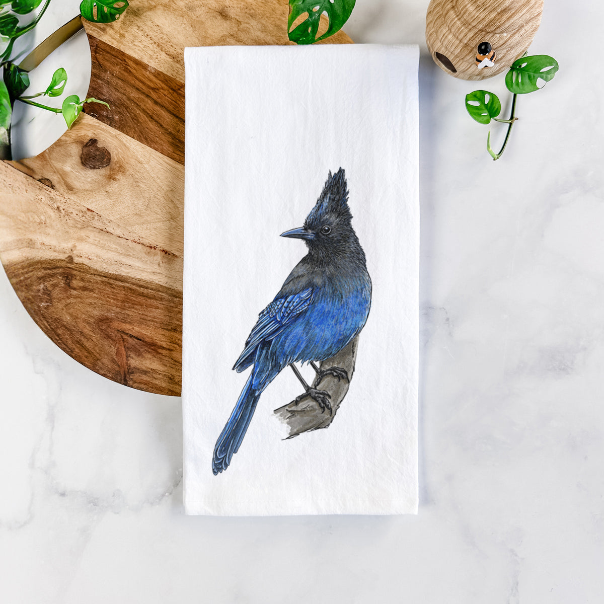 White cotton tea towel printed with a full-color illustration of a Steller's jay perched with its dark crest raised, on a marble counter beside a round wooden cutting board and greenery.