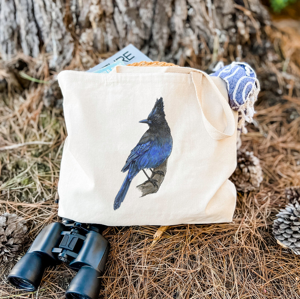Canvas tote bag printed with a full-color illustration of a Steller's jay perched with its dark crest raised, resting on dry grass beside a pair of binoculars.