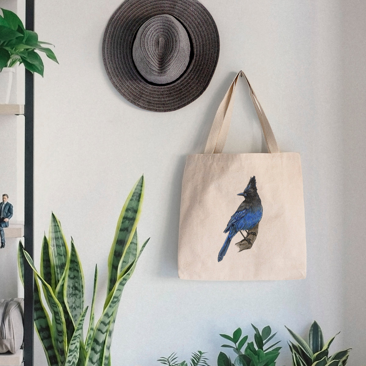 Canvas tote bag printed with a full-color illustration of a Steller's jay perched with its dark crest raised, hanging on a wall hook below a hat, next to potted plants.