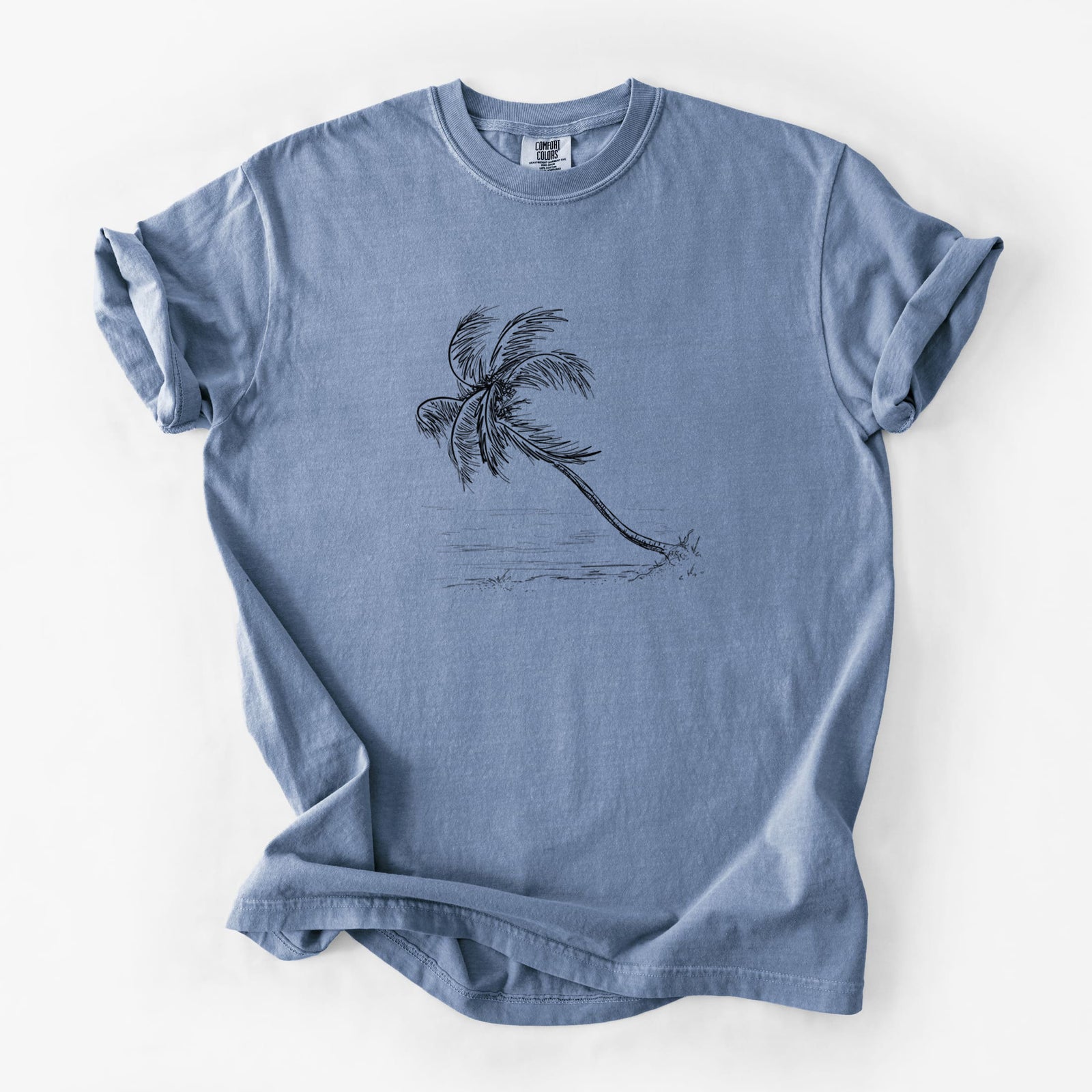 Blue jean Comfort Colors t-shirt laid flat, printed with a black ink illustration of a coconut palm leaning over a sandy beach.