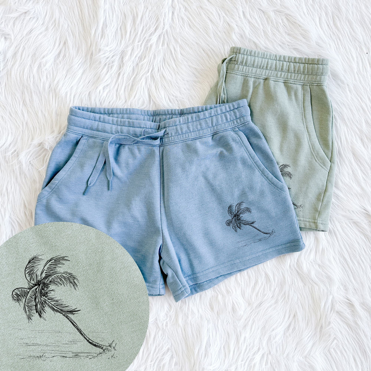 Two pairs of Cali Wave shorts in different colors printed with a black ink illustration of a coconut palm leaning over a sandy beach, with a close-up of the print.