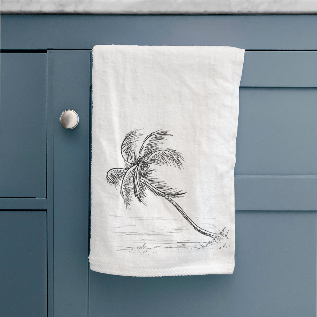 White hand towel printed with a black ink illustration of a coconut palm leaning over a sandy beach, hanging on a cabinet door.