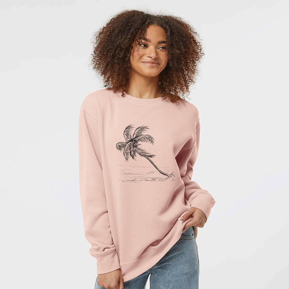 Model wearing a dusty pink pigment dyed crewneck sweatshirt printed with a black ink illustration of a coconut palm leaning over a sandy beach.