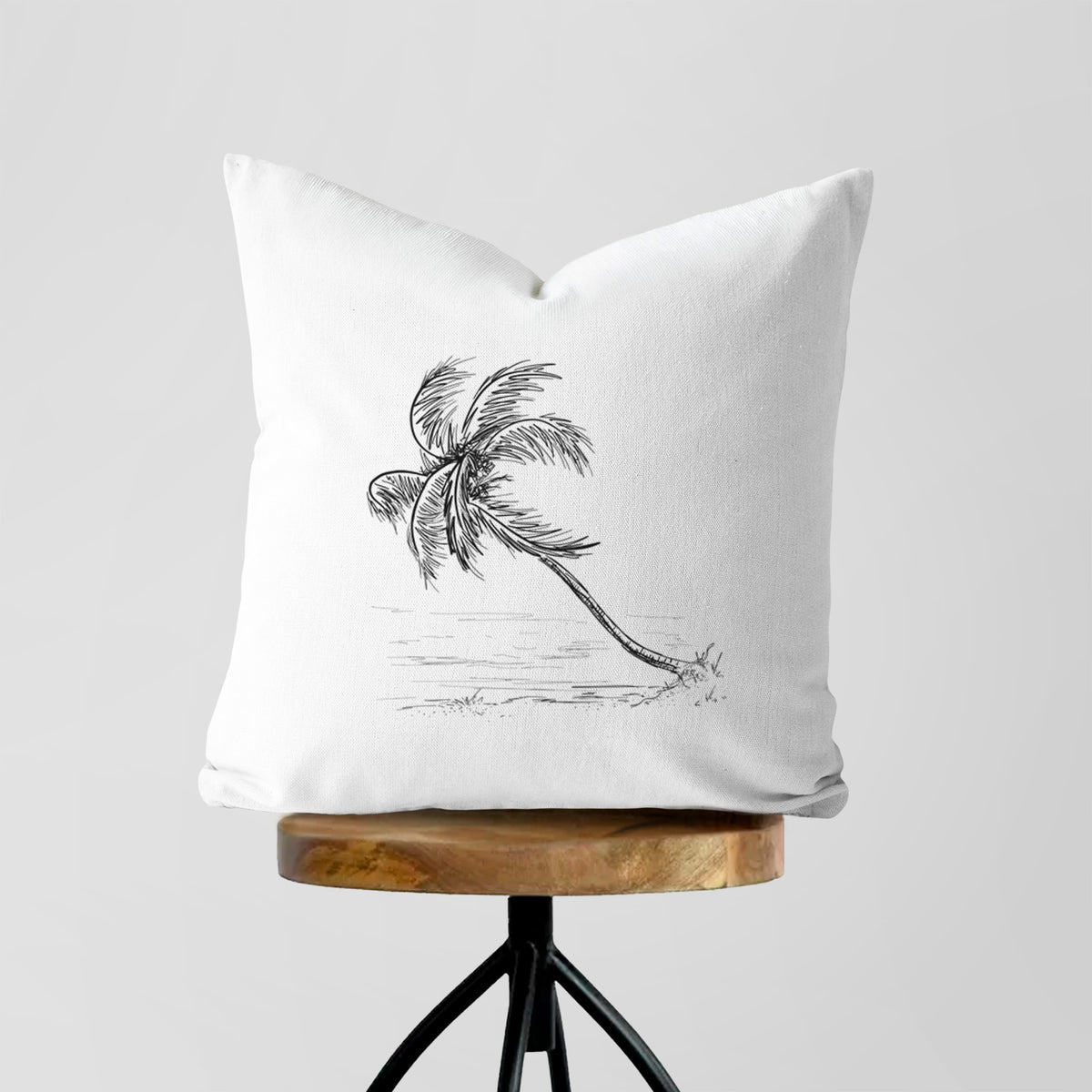 White pillow cover printed with a black ink illustration of a coconut palm leaning over a sandy beach, on a wooden stool.