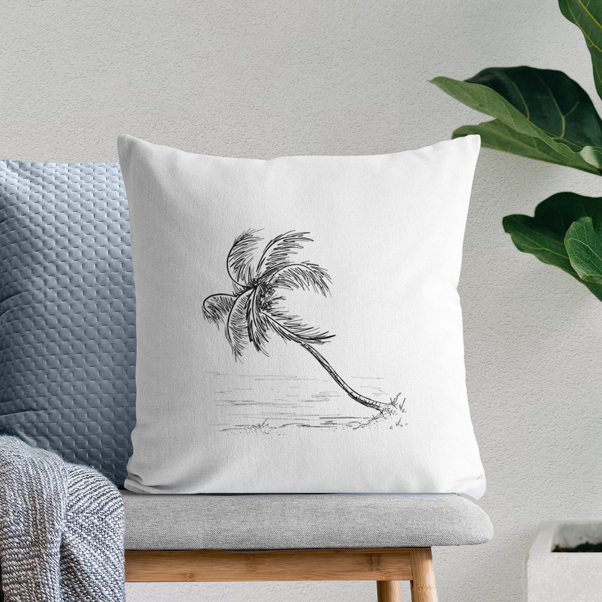 White pillow cover printed with a black ink illustration of a coconut palm leaning over a sandy beach, on a bench beside plants.