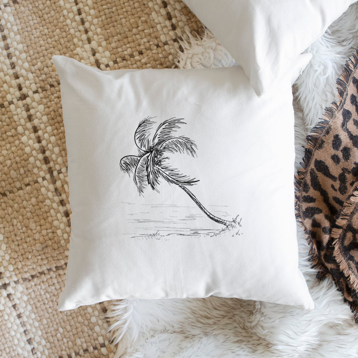 White pillow cover printed with a black ink illustration of a coconut palm leaning over a sandy beach, on a woven chair.