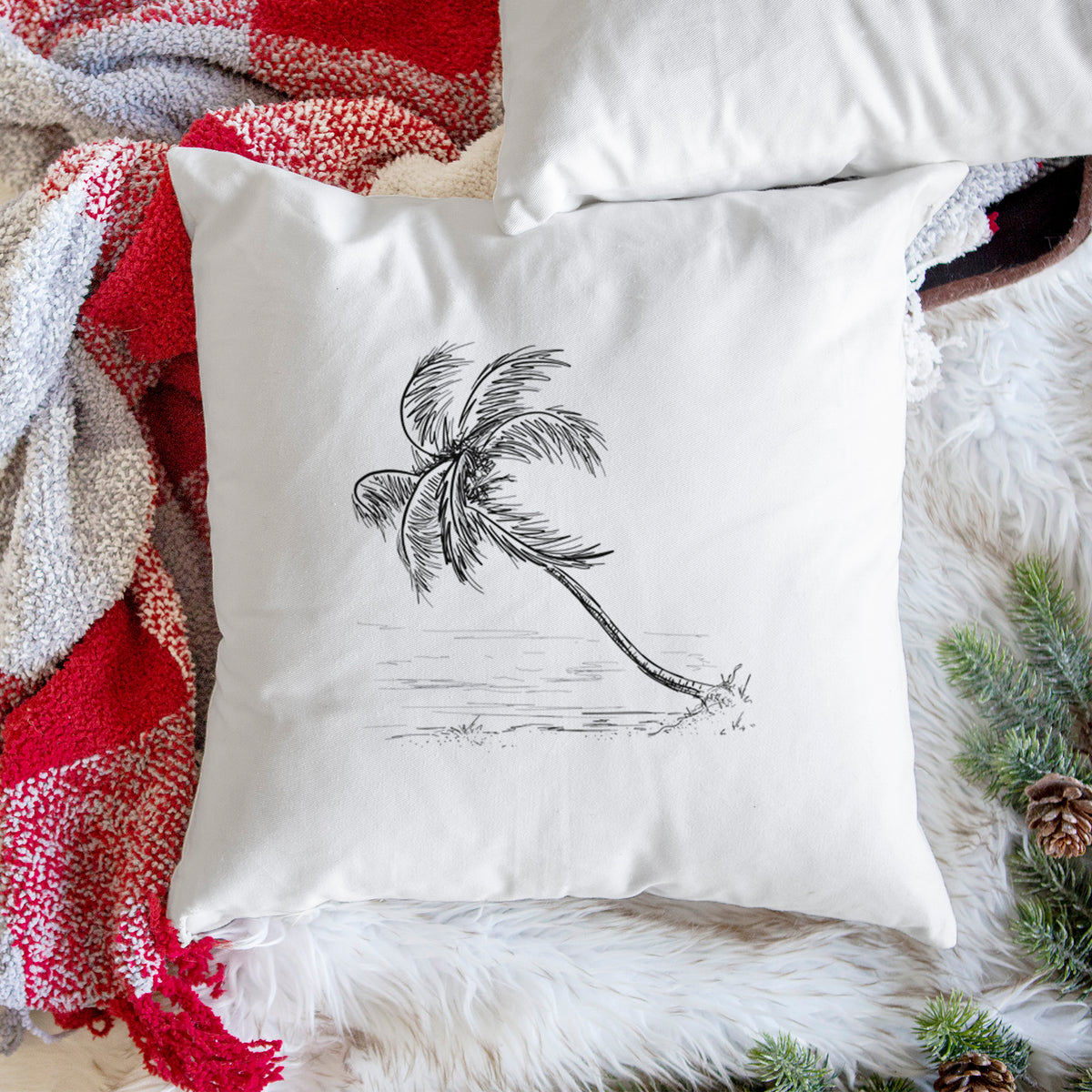 White pillow cover printed with a black ink illustration of a coconut palm leaning over a sandy beach, on a woven chair.